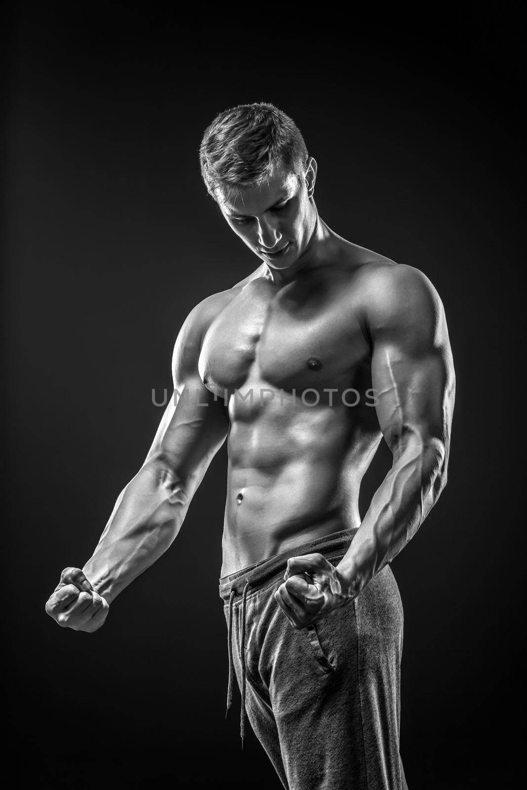 Image of very muscular man posing with naked torso in studio on black background. Black and white