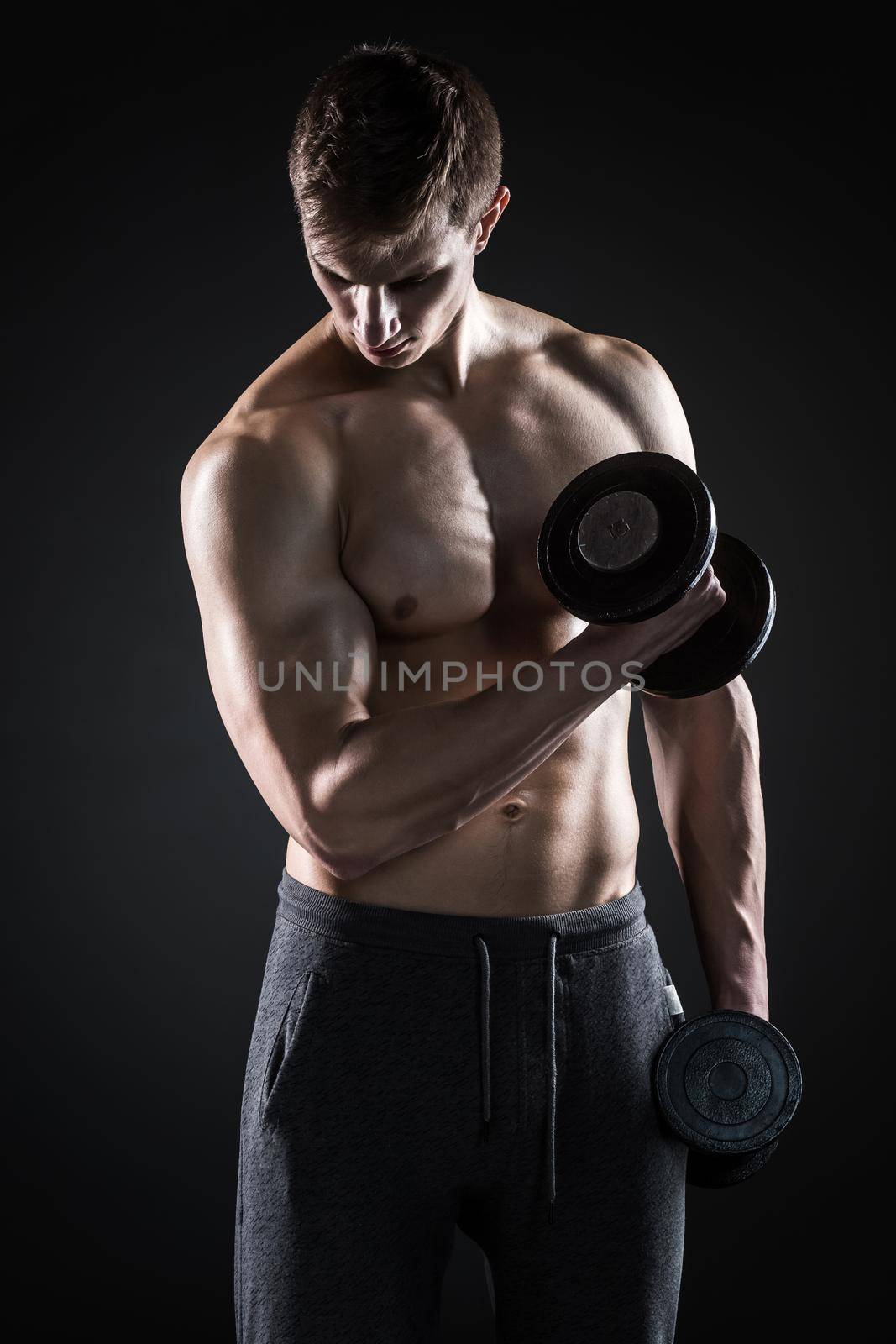 Young handsome man with naked torso and dumbbells in hand on black background