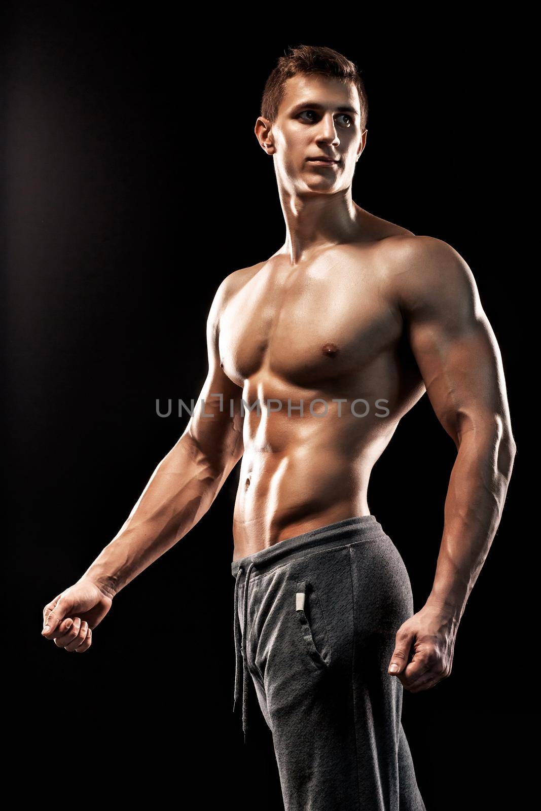 Image of very muscular man posing with naked torso in studio and looking behind on black background.