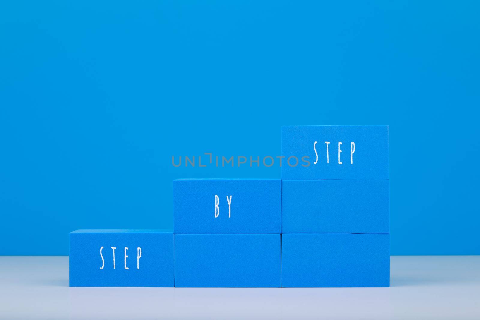 Ladder made of blue toy blocks with written step by step text on white table against blue background with copy space. Concept of business or personal development, success or achieving goals