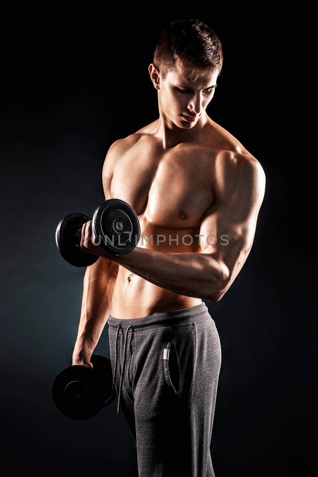 Athletic man showing muscular body and doing exercises with dumbbells on black background..