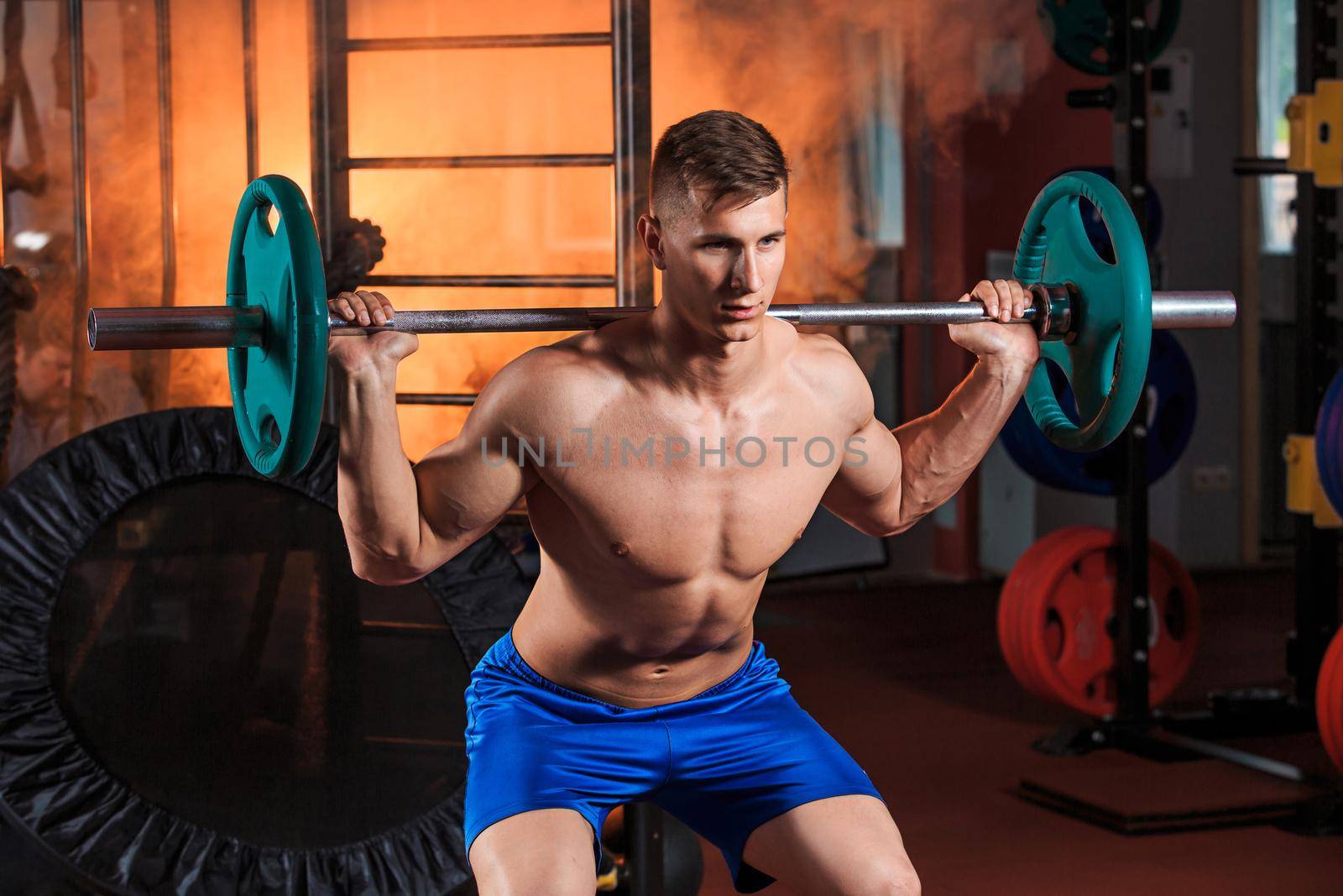 young man doing squats with barbell in the gym. Orange smoke on background