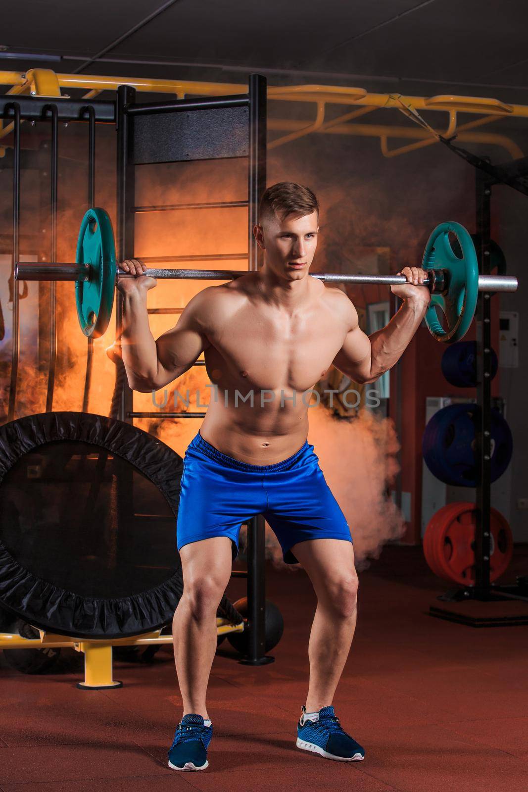 young man doing squats with barbell in the gym. Orange smoke on background