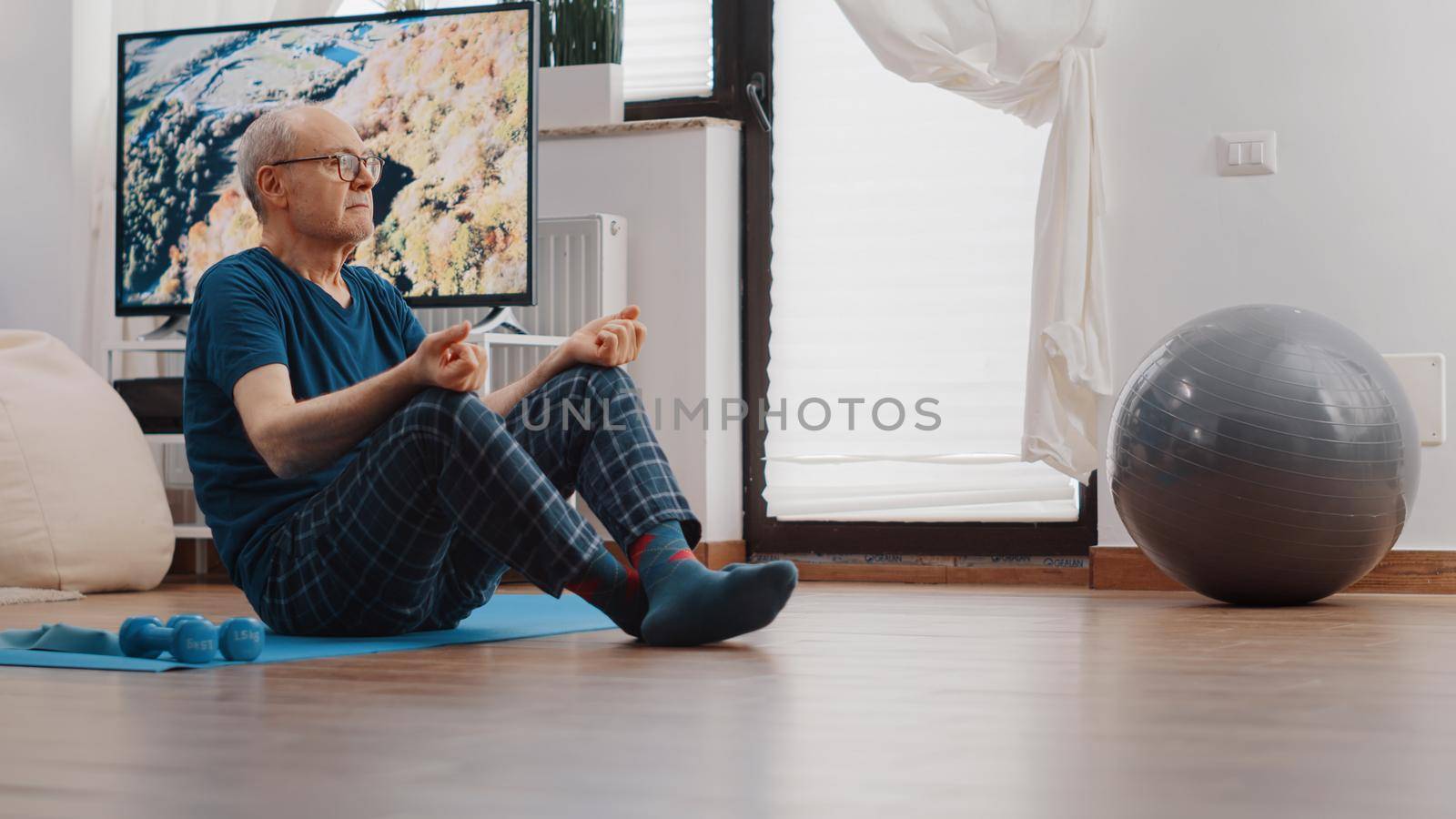 Senior man sitting in lotus position and doing meditation on yoga mat. Zen pensioner breathing to meditate and relax after workout exercise. Old person meditating for wellness and recreation