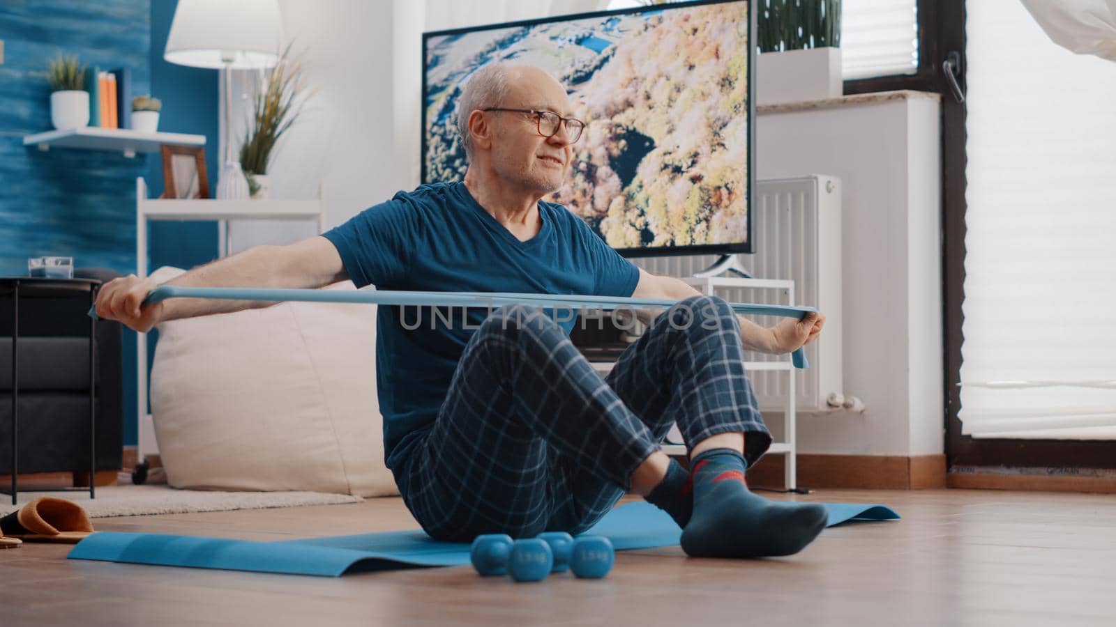 Old man using resistance band to stretch and pull, sitting on yoga mat. Senior person training with elastic belt to do arms muscles exercise at home. Elder adult doing physical activity