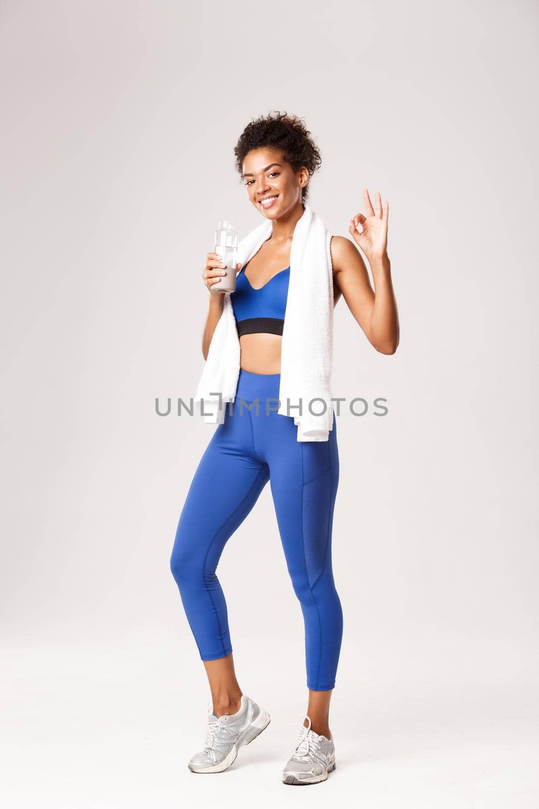 Full length of attractive and fit african-american fitness girl, standing with towel and water bottle, showing okay sign and smiling satisfied after good workout, white background.