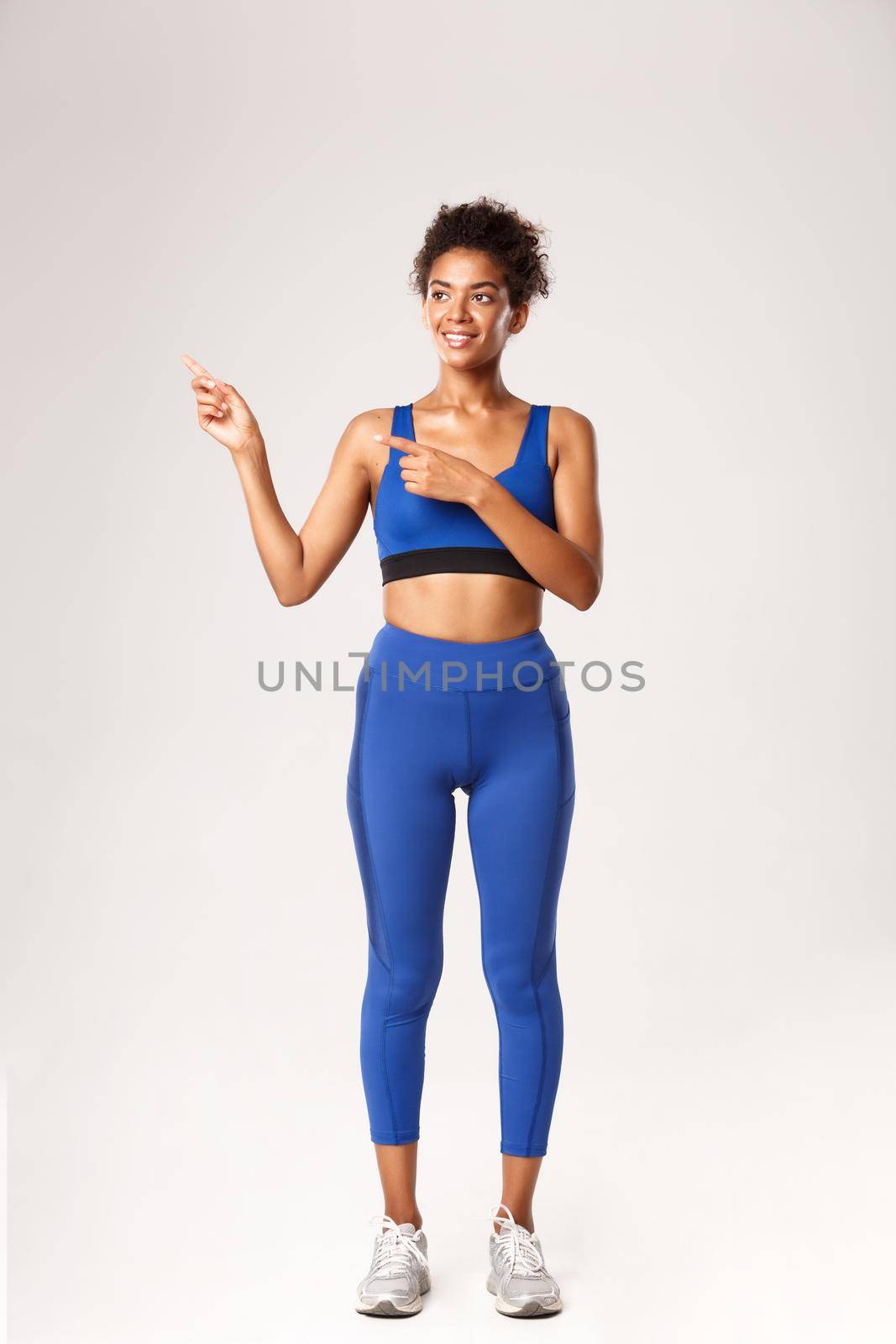 Full length of athletic african-american gitness girl in blue sportswear, pointing fingers left and looking pleased, recommend workout equipment, standing over white background by Benzoix