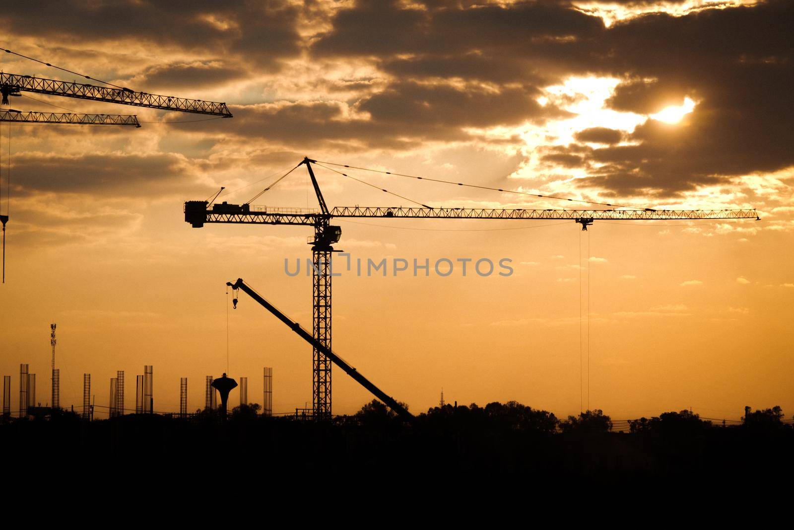 Silhouette of tower crane and large buildings construction site at sunset in evening time. New construction site with cranes on sunset background. by TEERASAK