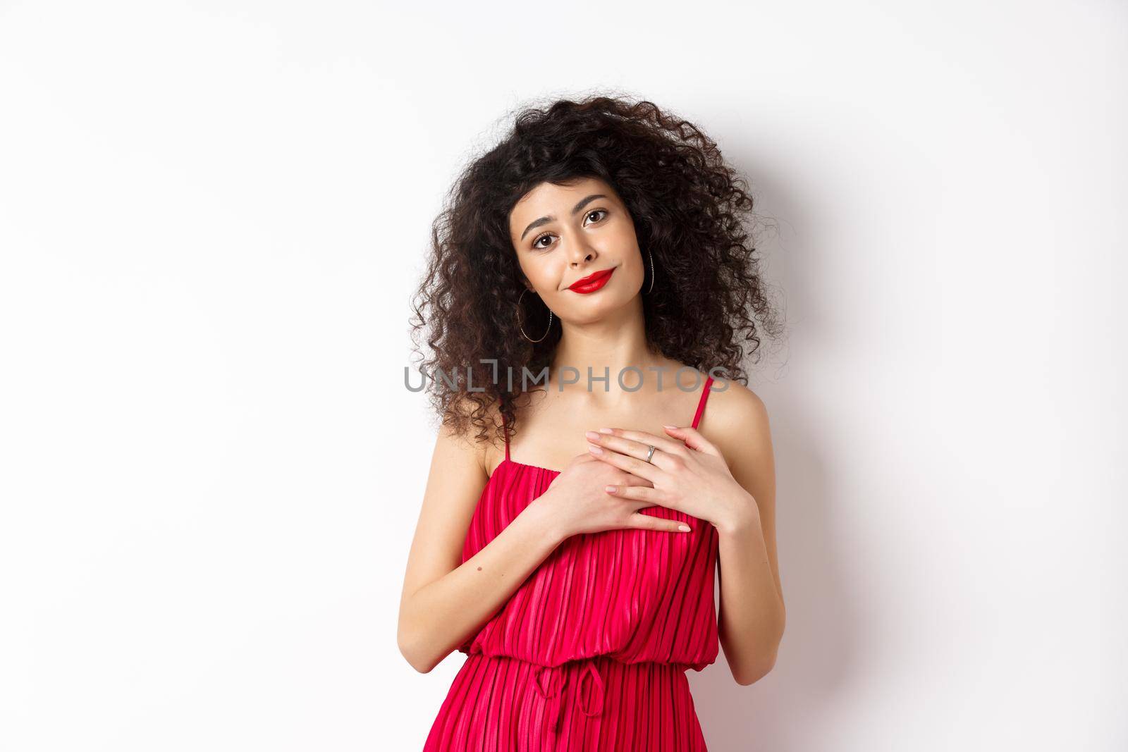 Touched young woman with curly hair, wearing red dress, holding hands on heart and smiling grateful, say thank you, standing over white background.