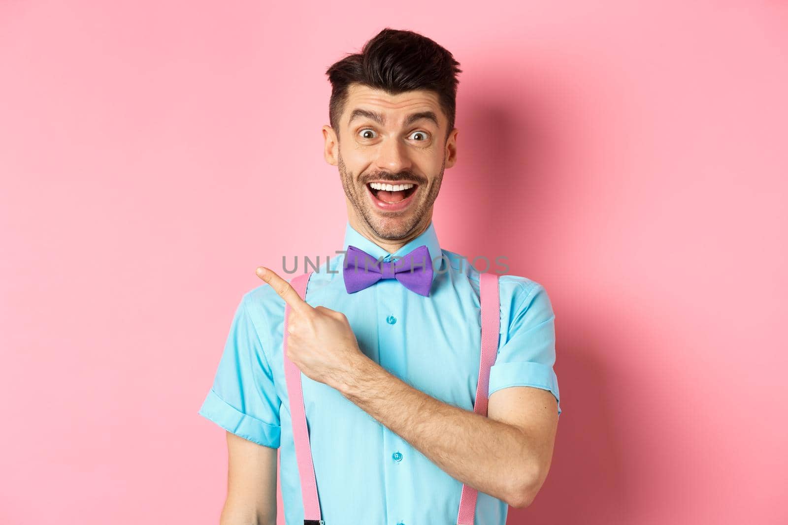 Happy young man checking out promo offer, smiling amazed and pointing finger left at copy space, standing near logo on pink background.