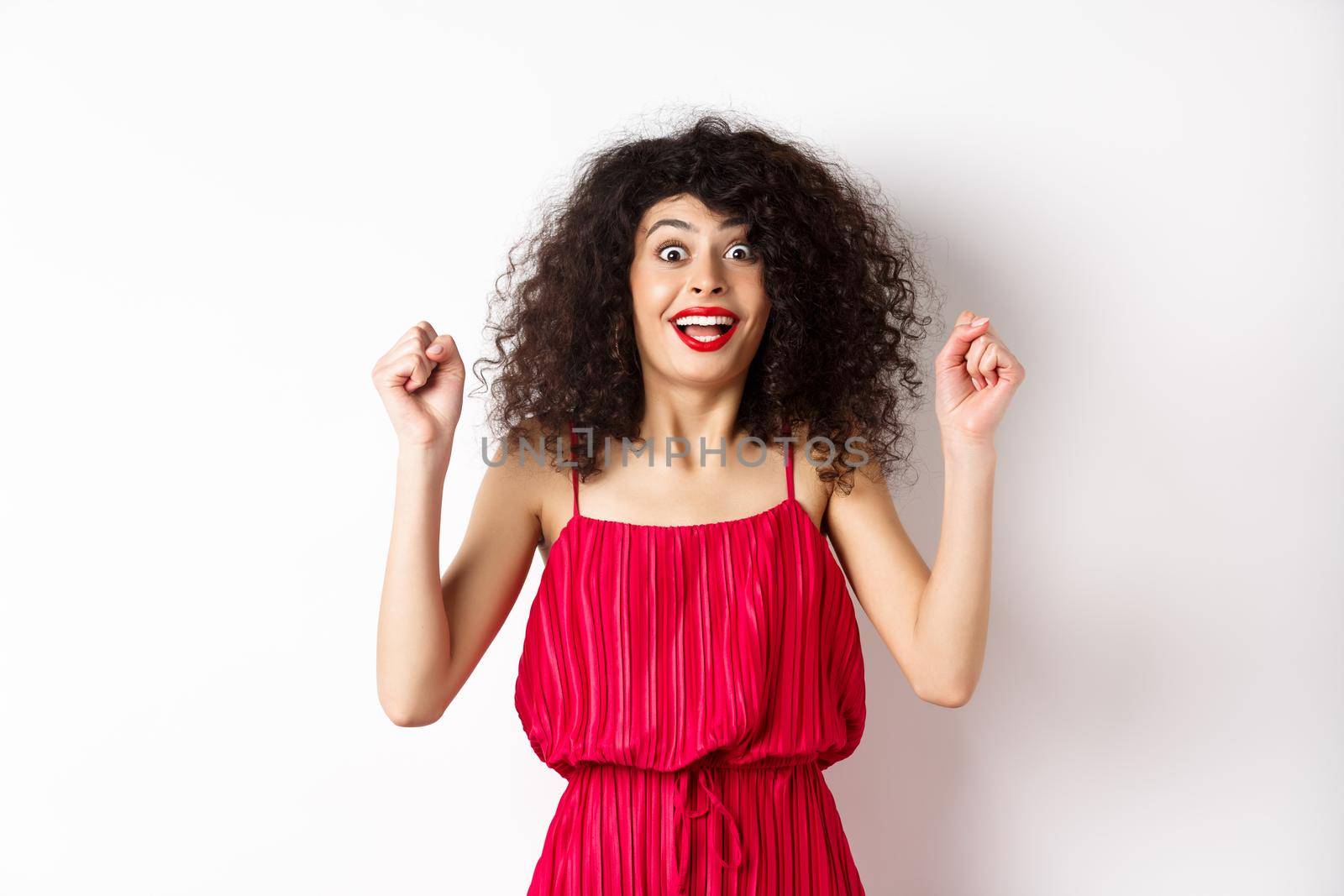 Excited curly-haired woman with evening makeup, gasping fascinated, raising hands up and rejoicing, winning prize and celebrating, standing against white background.