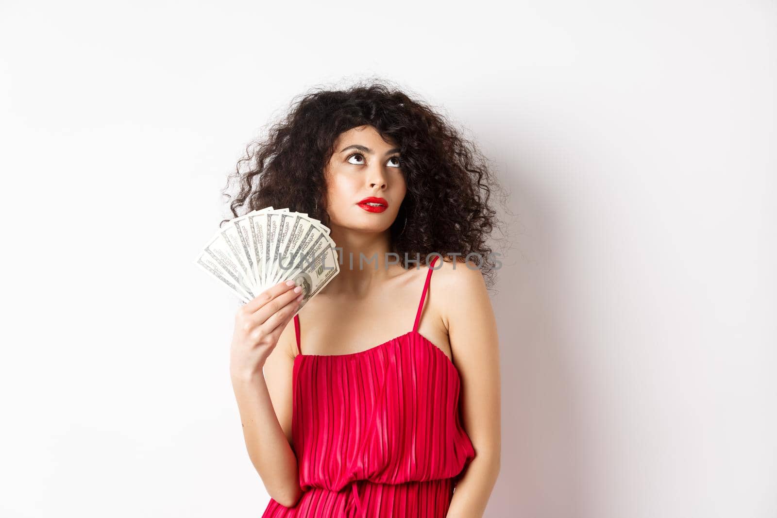 Elegant beautiful woman in red dress, waving with dollars and looking fabulous, standing on white background. Copy space