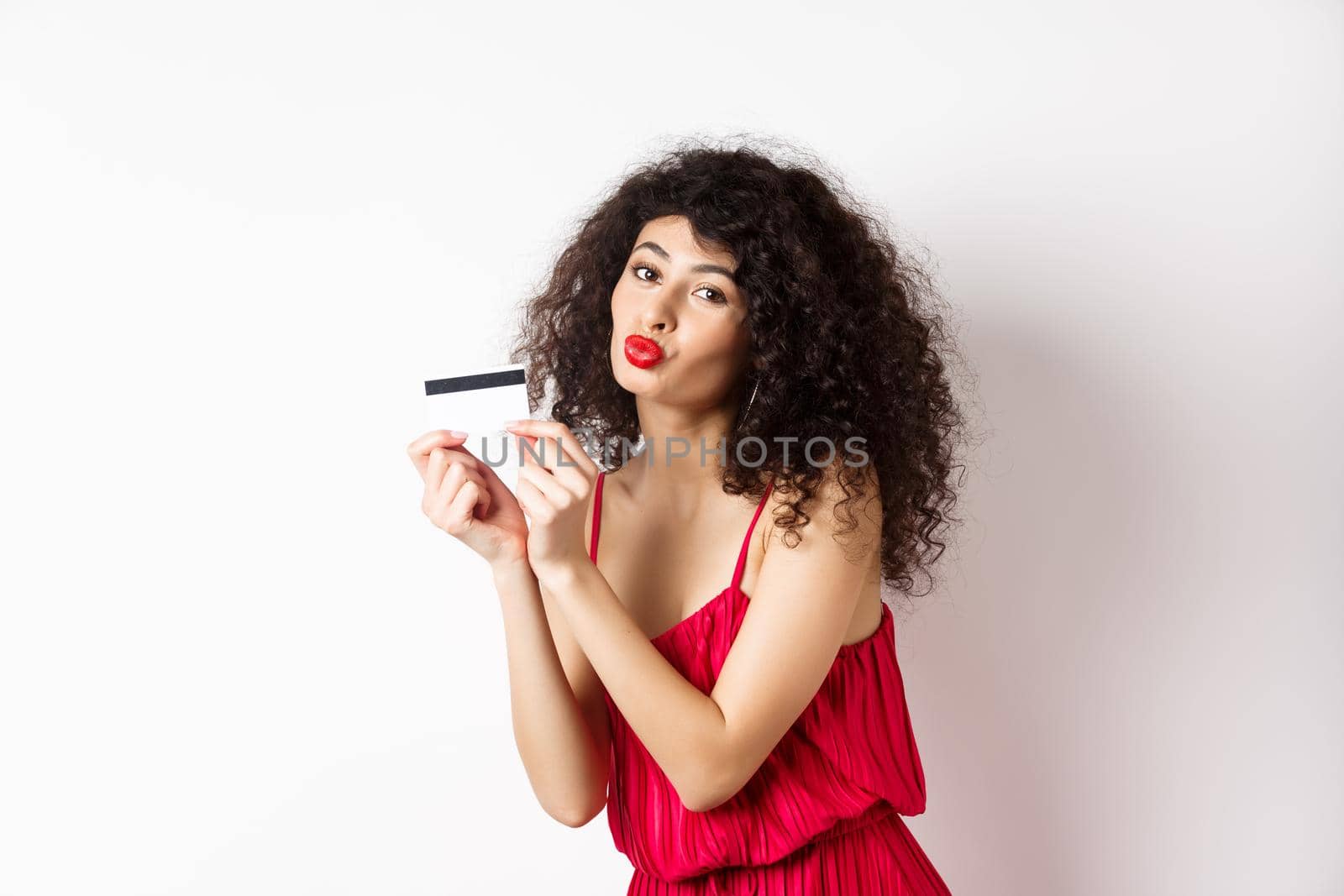 Shopping. Beautiful lady with curly hair, pucker lips, kissing plastic credit card, standing in red dress against white background.