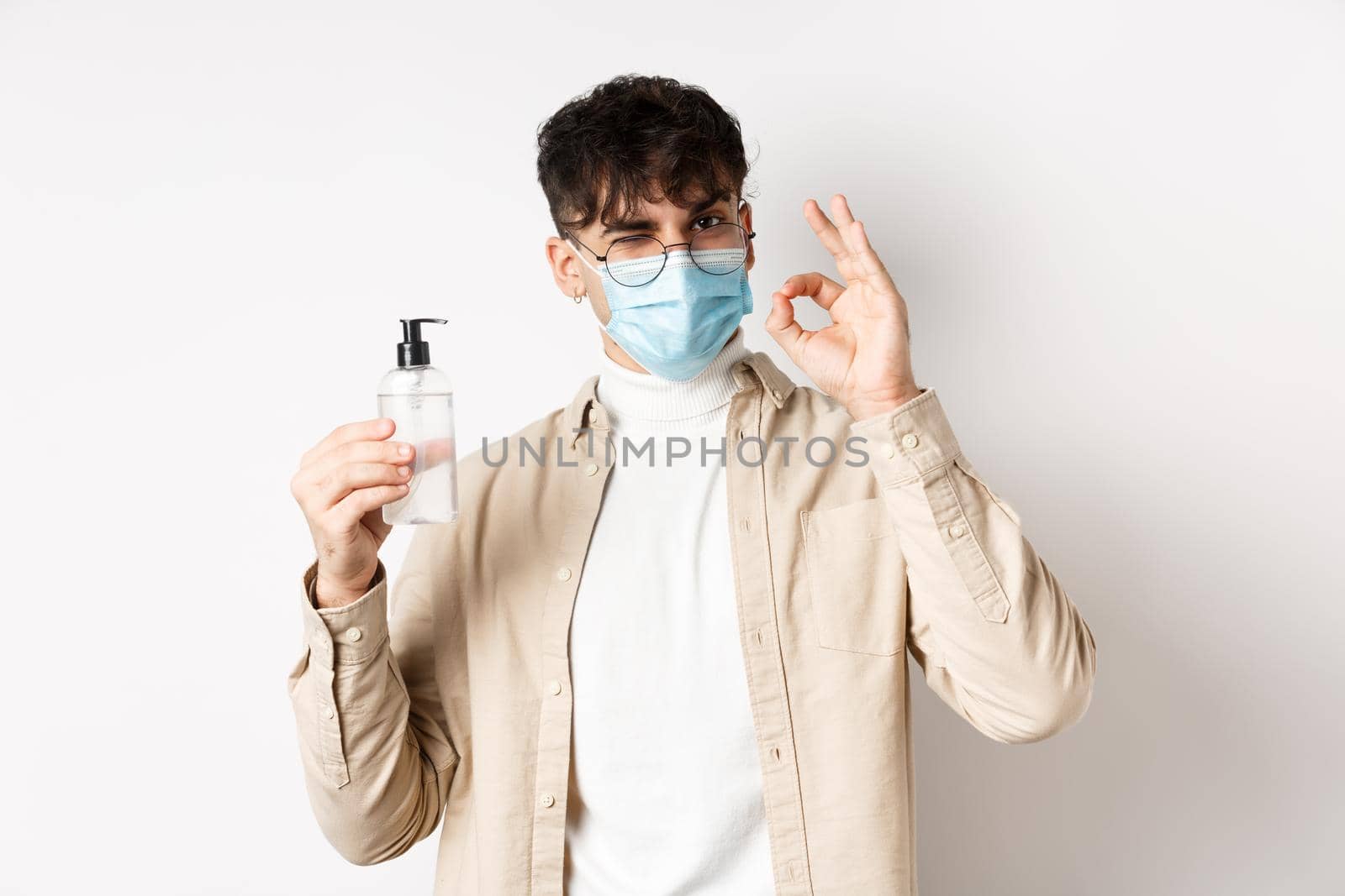 Health, covid and quarantine concept. Portrait of natural guy in glasses and face mask showing bottle of good hand sanitizer, make okay sign, recommending product, white background by Benzoix