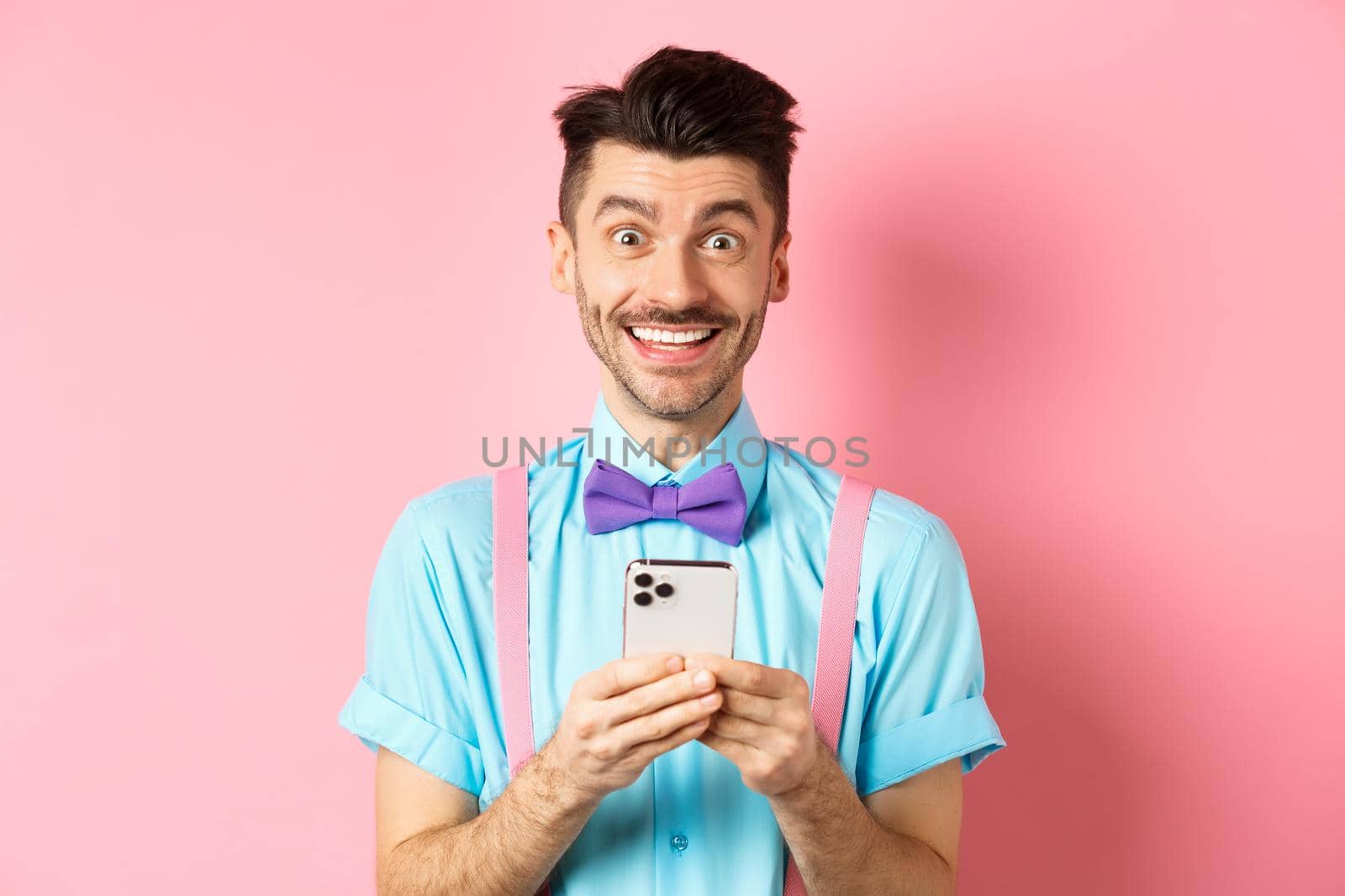 Online shopping. Happy guy in bow-tie, smiling at camera and using smartphone, standing on pink background by Benzoix