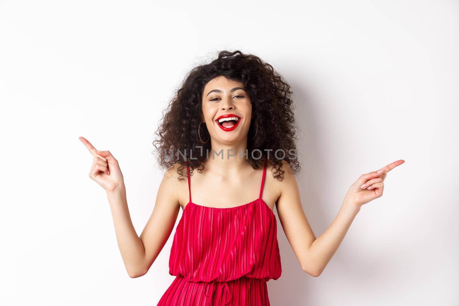 Happy beautiful lady in red dress, laughing and pointing sideways, showing two ways, standing on white background.