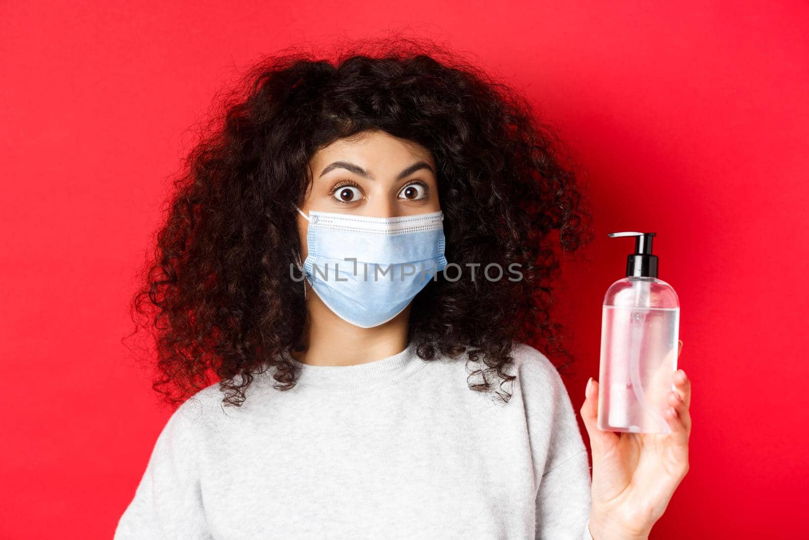 Covid-19, pandemic and quarantine concept. Excited girl with curly hair, wearing medical mask, showing bottle of hand sanitizer or antiseptic, standing on red background by Benzoix