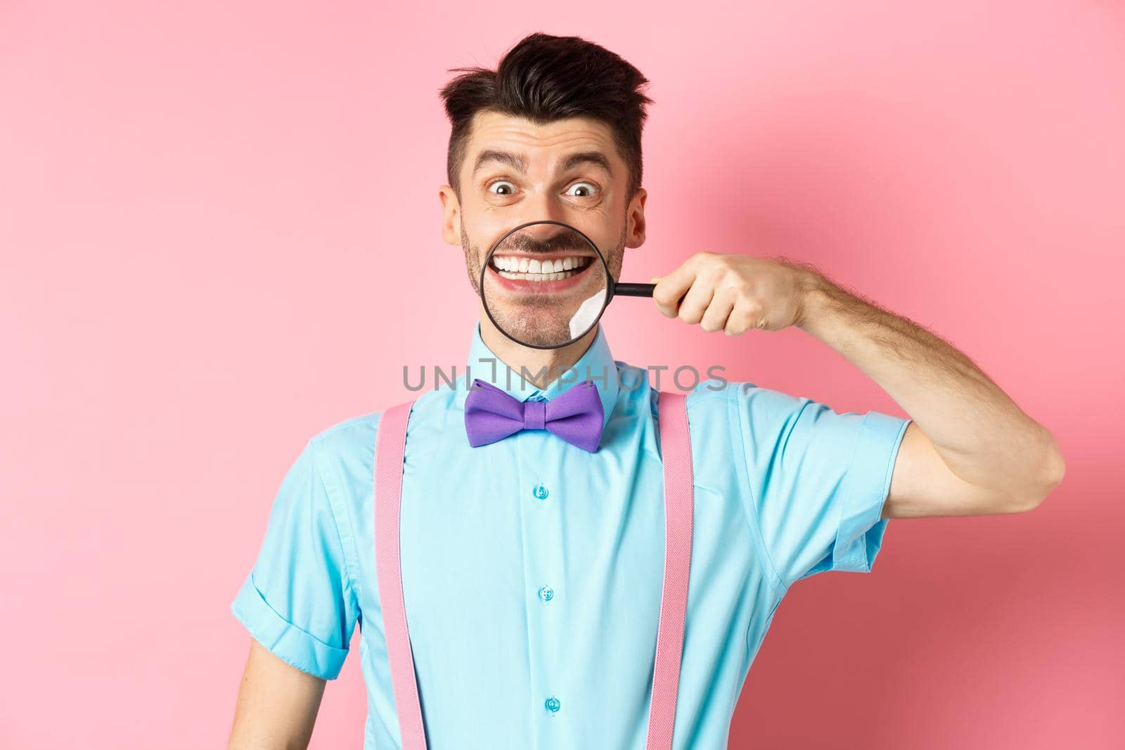 Funny caucasian guy in bow-tie showing his white smile teeth with magnifying glass, looking cheerful at camera, standing on pink background by Benzoix