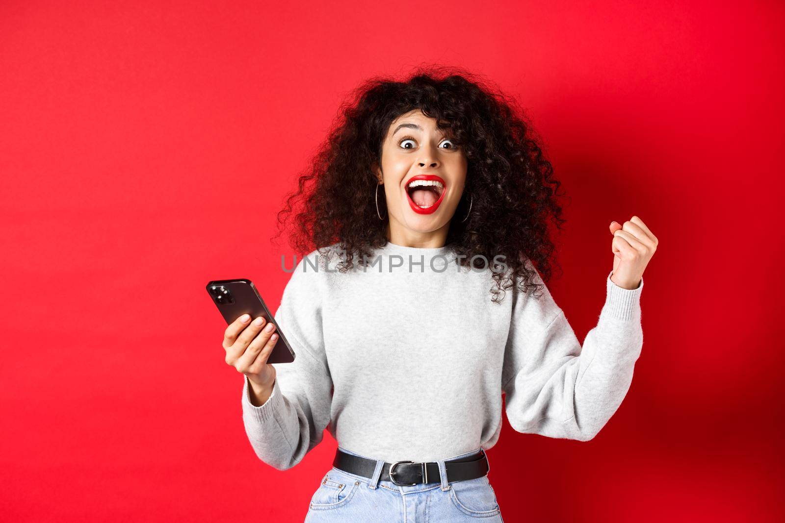 Excited caucasian woman with curly hair, chanting, winning online prize, holding smartphone and triumphing, standing on red background.