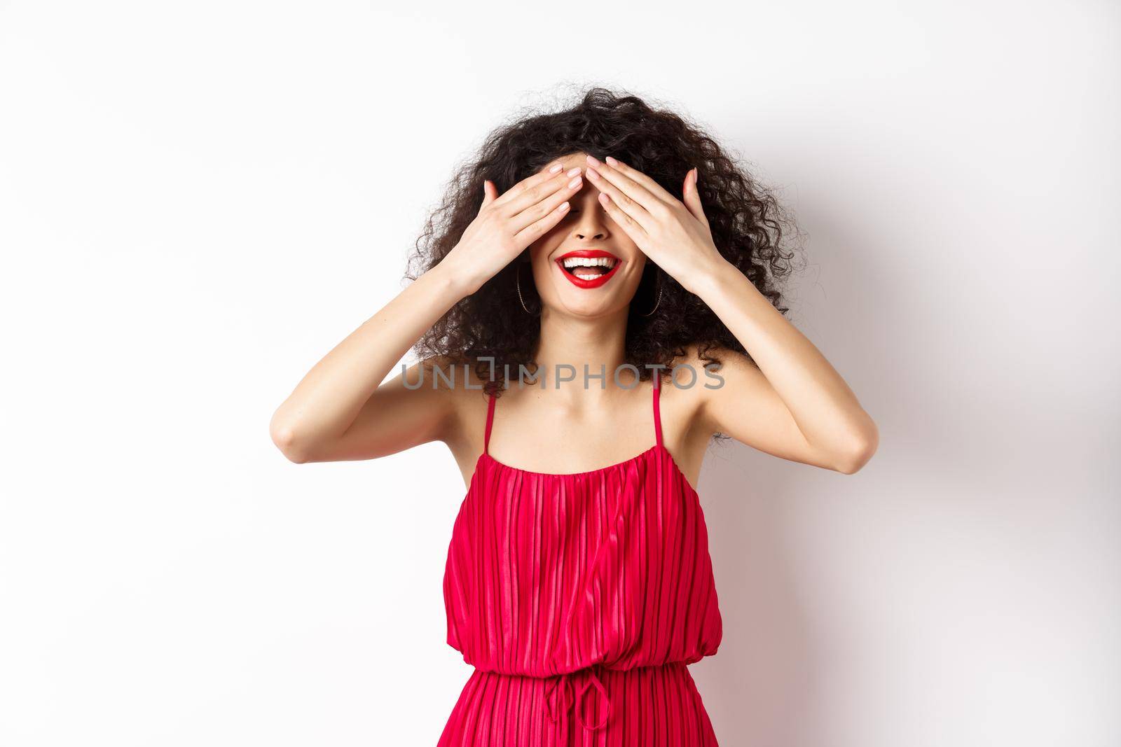 Excited curly-haired woman in party dress, close eyes and smiling with red lips, waiting for surprise with happy face, standing on white background.
