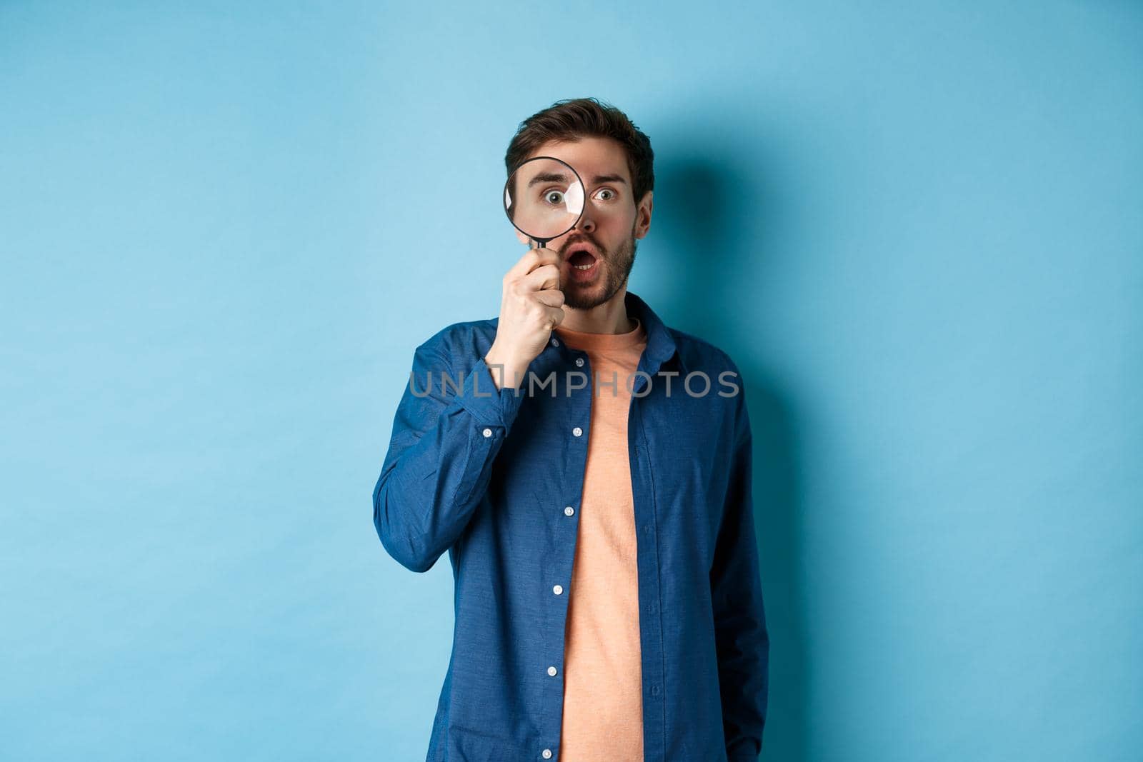 Impressed man look in awe through magnifying glass, drop jaw and stare at camera, standing on blue background.