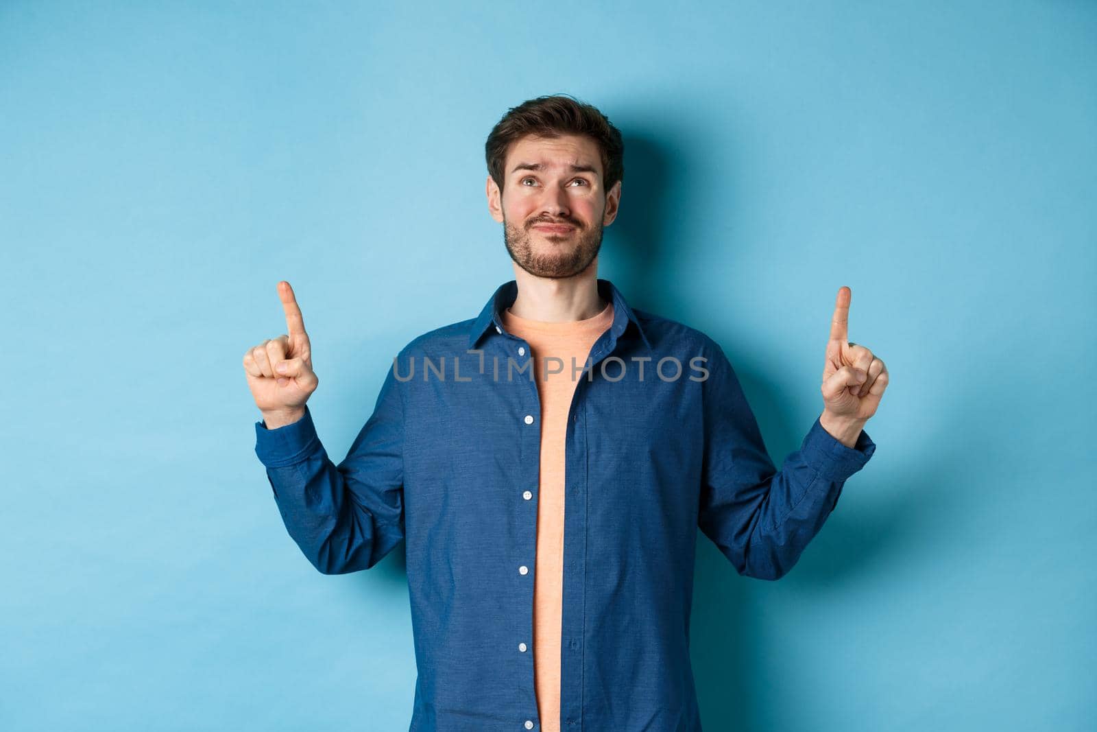 Skeptical young guy smirk and look doubtful, pointing fingers up upset, standing on blue background by Benzoix
