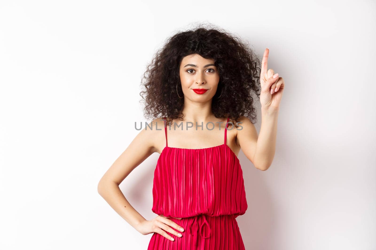 Smiling caucasian woman with curly hair, wearing red dress, showing rule number one gesture, raising finger and looking confident at camera, white background.
