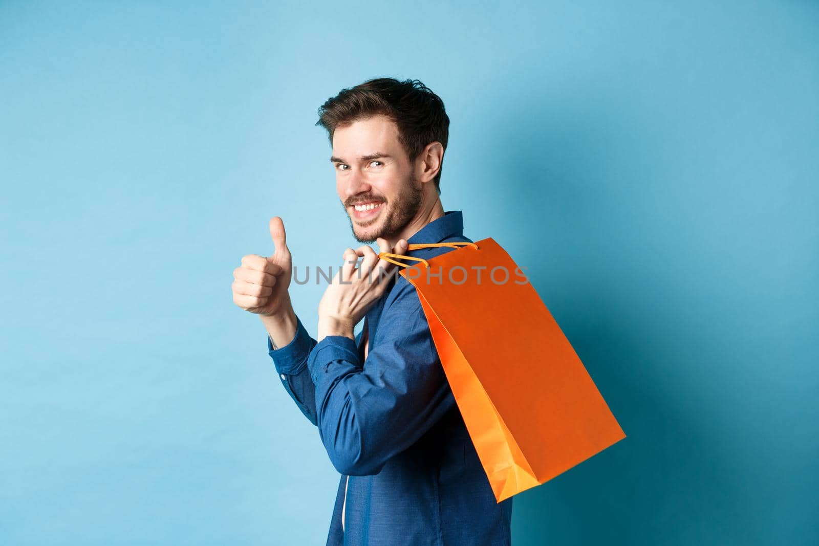 Cheerful shopper holding orange shopping bag on shoulder, turn around at camera with thumbs up, recommending store, blue background.