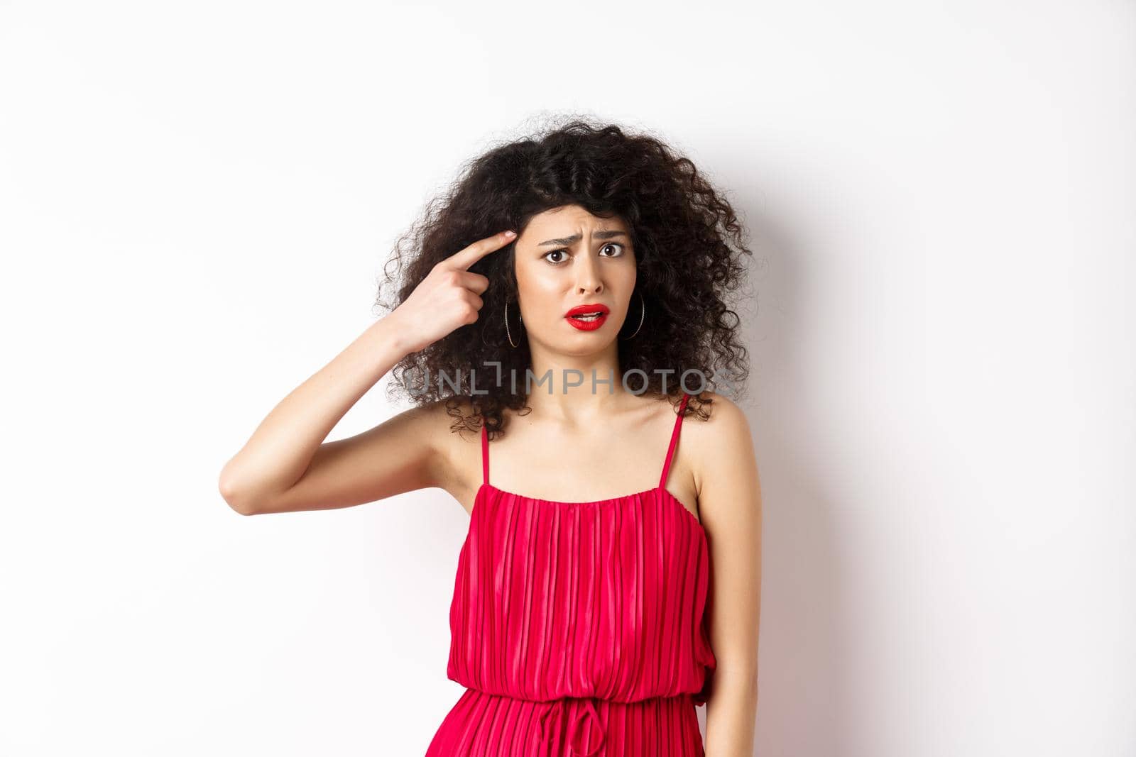 Shocked and confused caucasian woman in red dress scolding someone crazy, pointing finger at head and frowning, standing over white background.