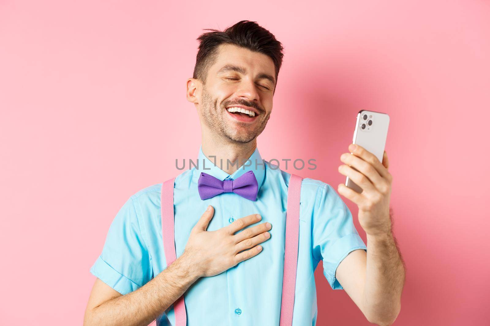 Technology concept. Happy caucasian man video chat and laugh at smartphone, standing over pink background.