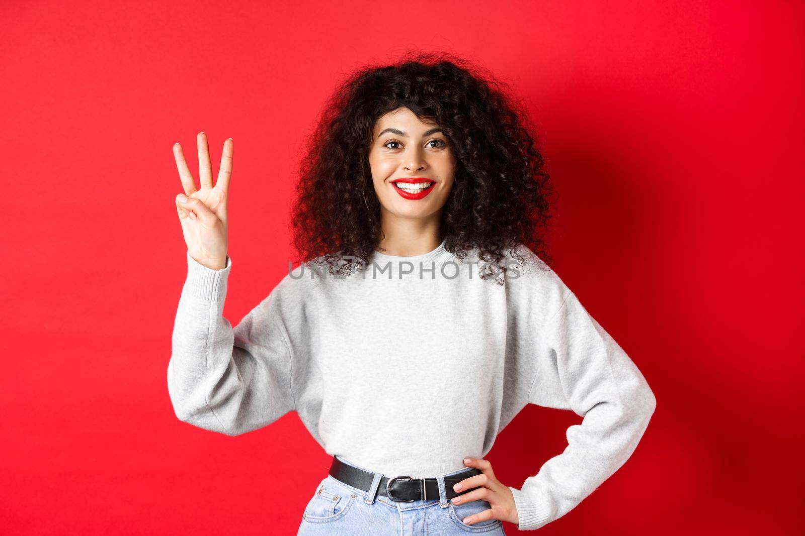 Cheerful female model showing number three and smiling, making an order, standing in sweatshirt on red background.