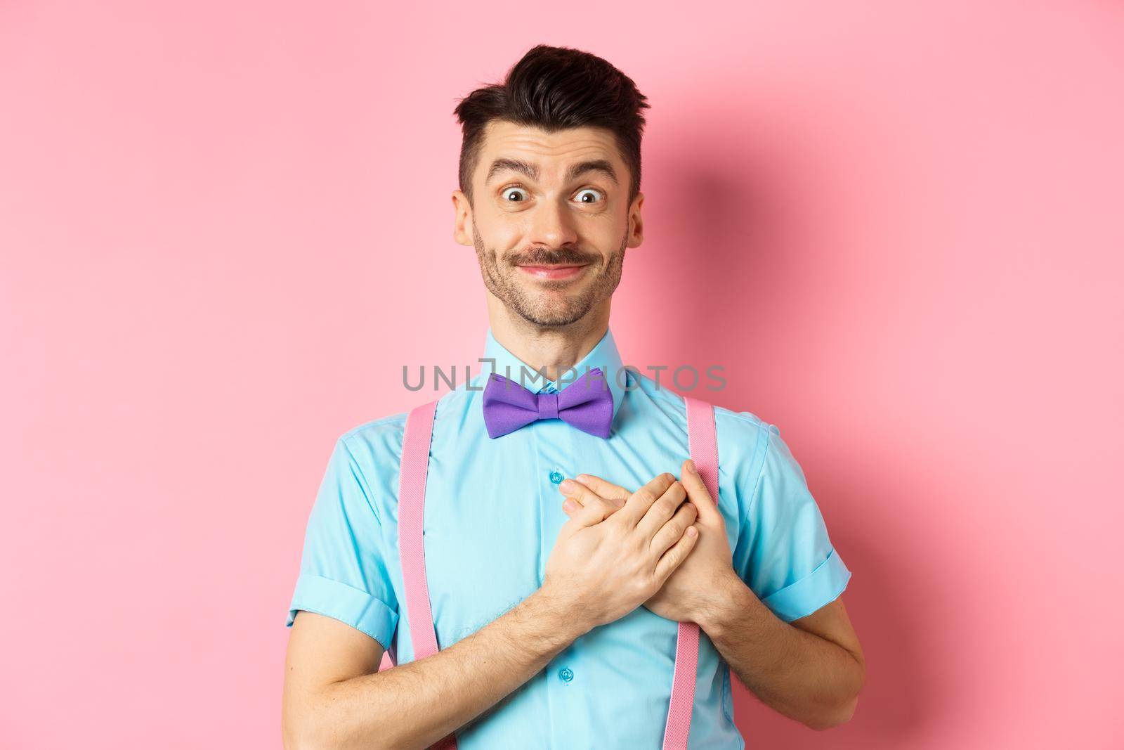 Cheerful young man with moustache, wearing shirt and bow-tie, holding hands on heart and smiling grateful, saying thank you, standing on pink background.