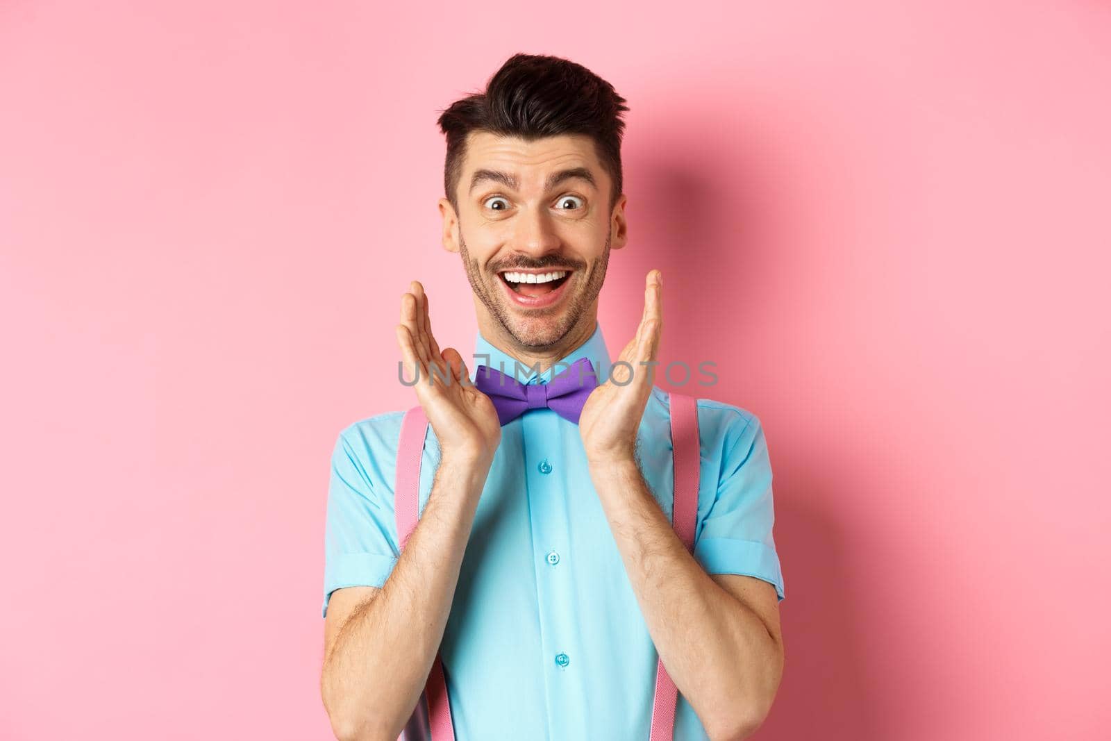 Happy young man looking with amazement and disbelief, smiling at camera, holding hands near face, standing in bow-tie and shirt on pink background.