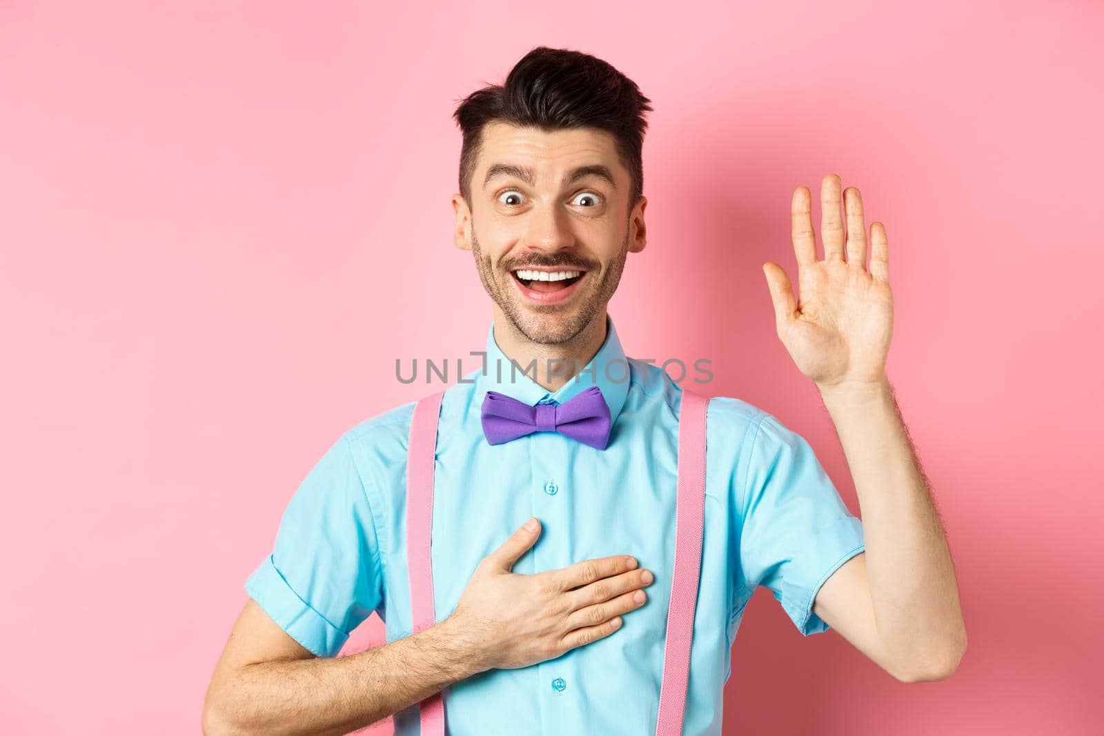 Happy young man telling truth, making promise, holding hand on heart and arm raised, swearing to be honest, smiling at camera, standing over pink background.