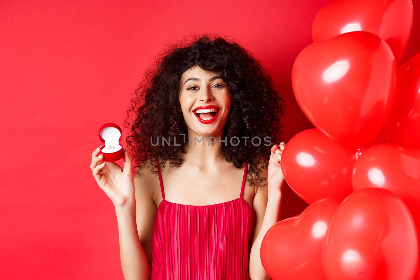 Happy beautiful lady feeling excited about marriage proposal, showing engagement ring and laughing, standing near heart balloons on red background.