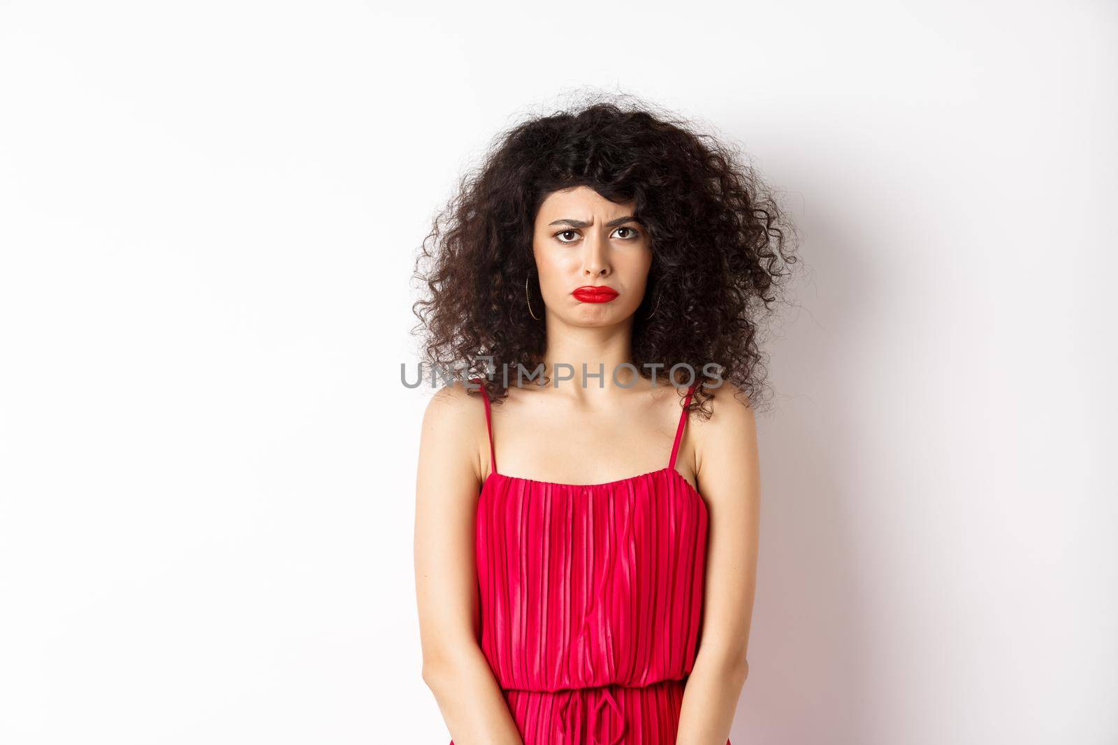Sad frowning lady in red dress complaining, looking jealous or lonely, standing upset over white background. Copy space