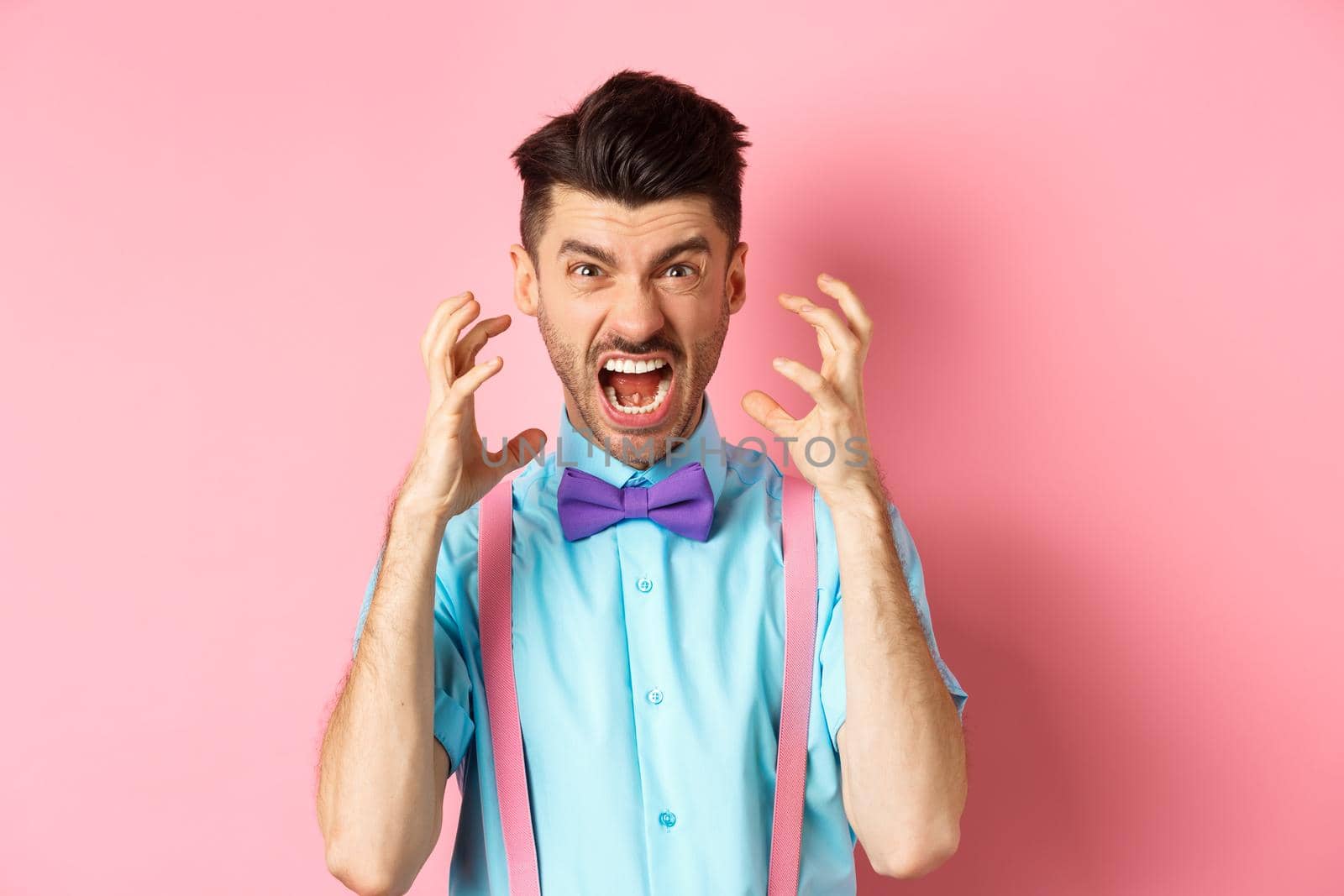 Frustrated young man losing temper, screaming and looking angry at camera, shaking hands pissed-off, standing mad on pink background.