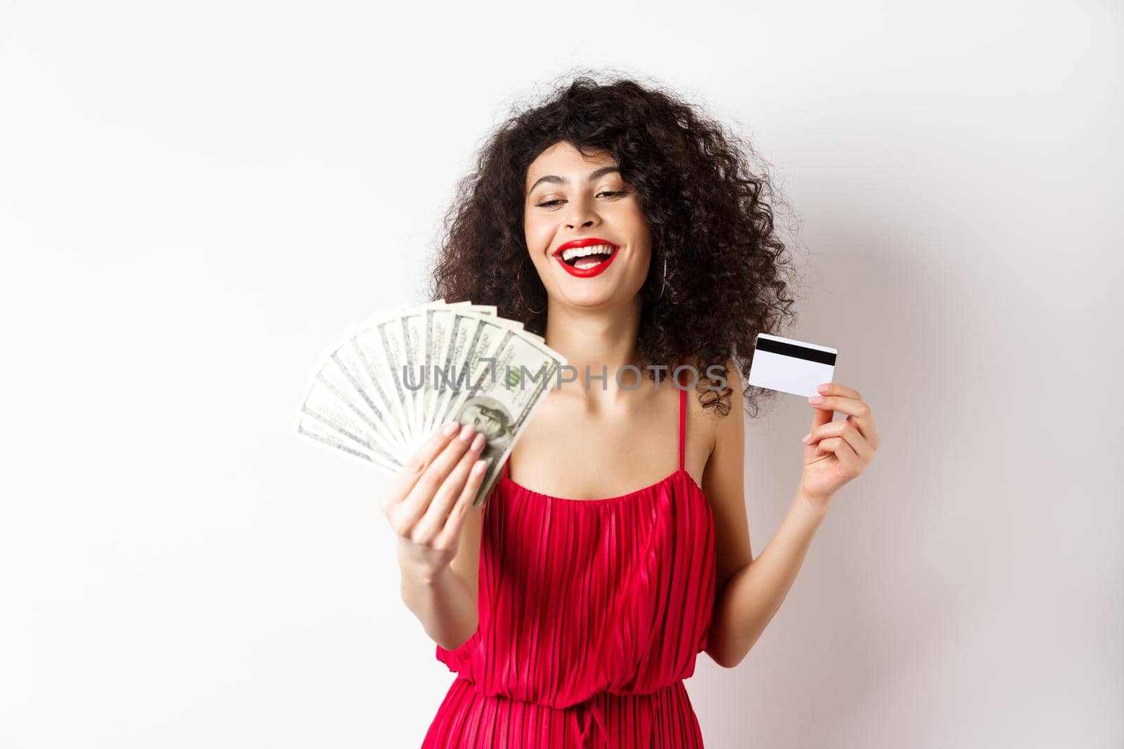 Shopping. Rich successful woman with curly hair and red dress, holding plastic credit card and looking pleased at dollar bills, white background.