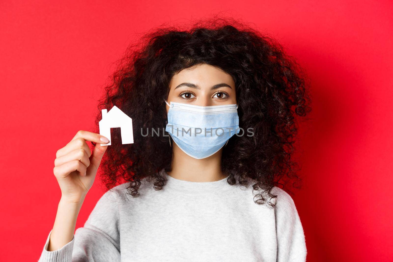 Real estate and covid-19 concept. Excited woman in medical mask showing small paper house cutout, standing on red background.