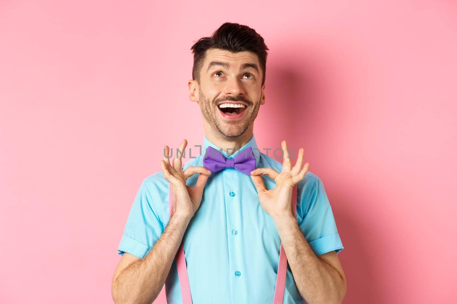 Happy young man with moustache, touching his bow-tie and laughing, looking up at logo, standing on pink background by Benzoix