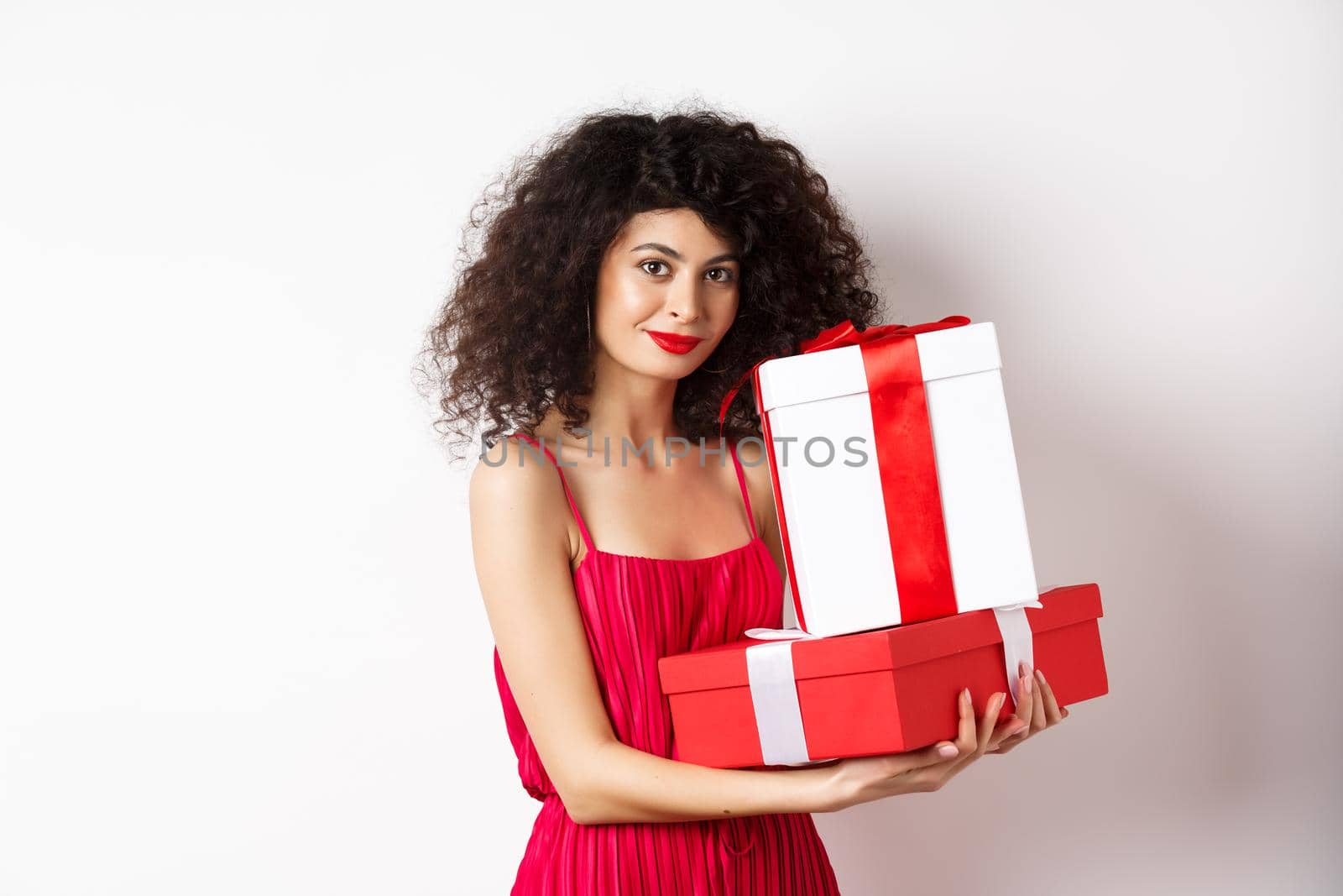 Elegant woman holding Valentines day gifts and smiling romantic at camera, standing in red dress on white background.