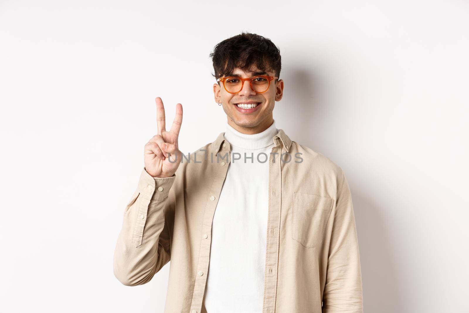 Handsome caucasian guy in glasses showing two fingers and smiling, standing on white background.