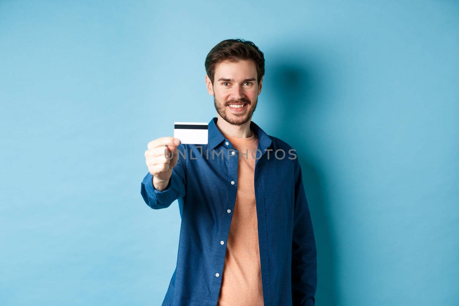Cheerful caucasian man showing plastic credit card and smiling, recommending bank, standing on blue background.