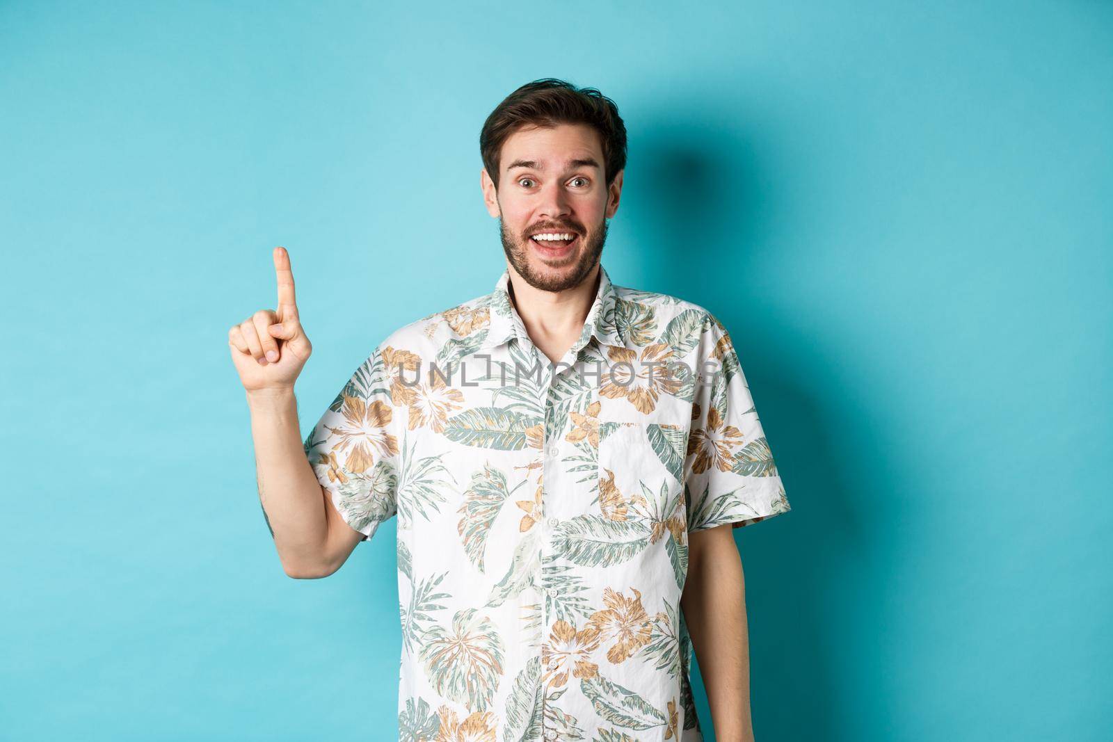 Excited guy in hawaiian shirt talking about vacation, looking amazed and pointing up, standing on blue background.