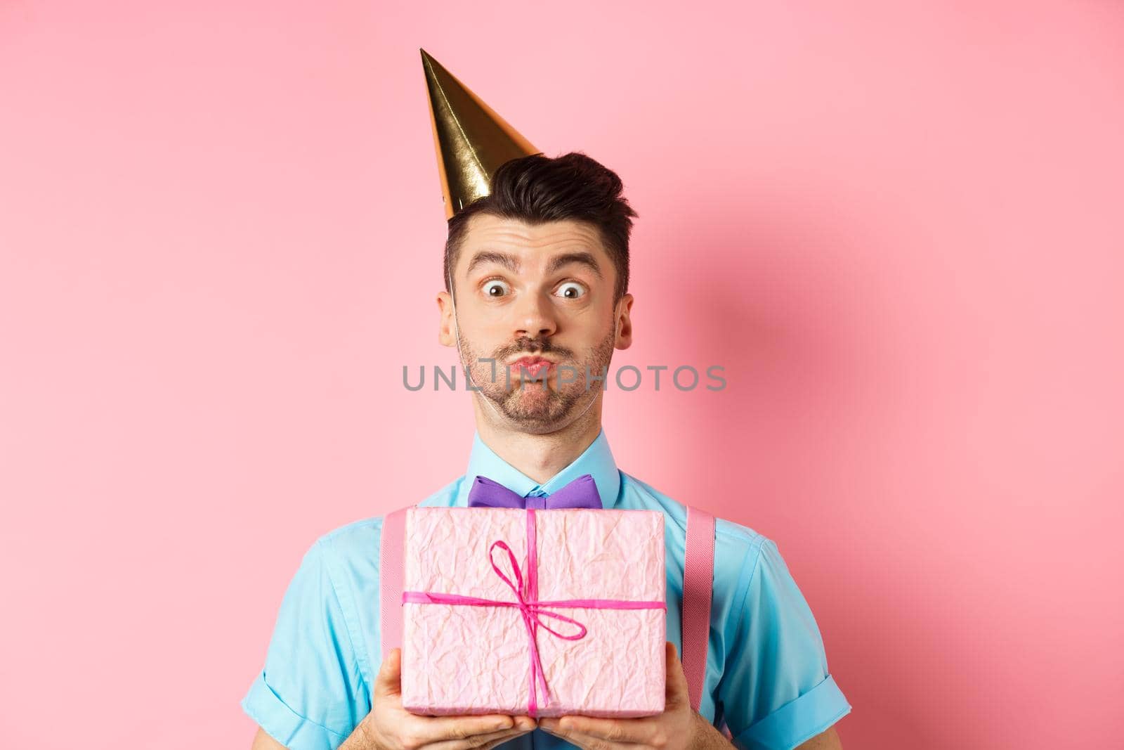 Holidays and celebration concept. Funny guy staring at camera surprised, wearing party hat, holding birthday gift and holding breath, pouting at camera, pink background.