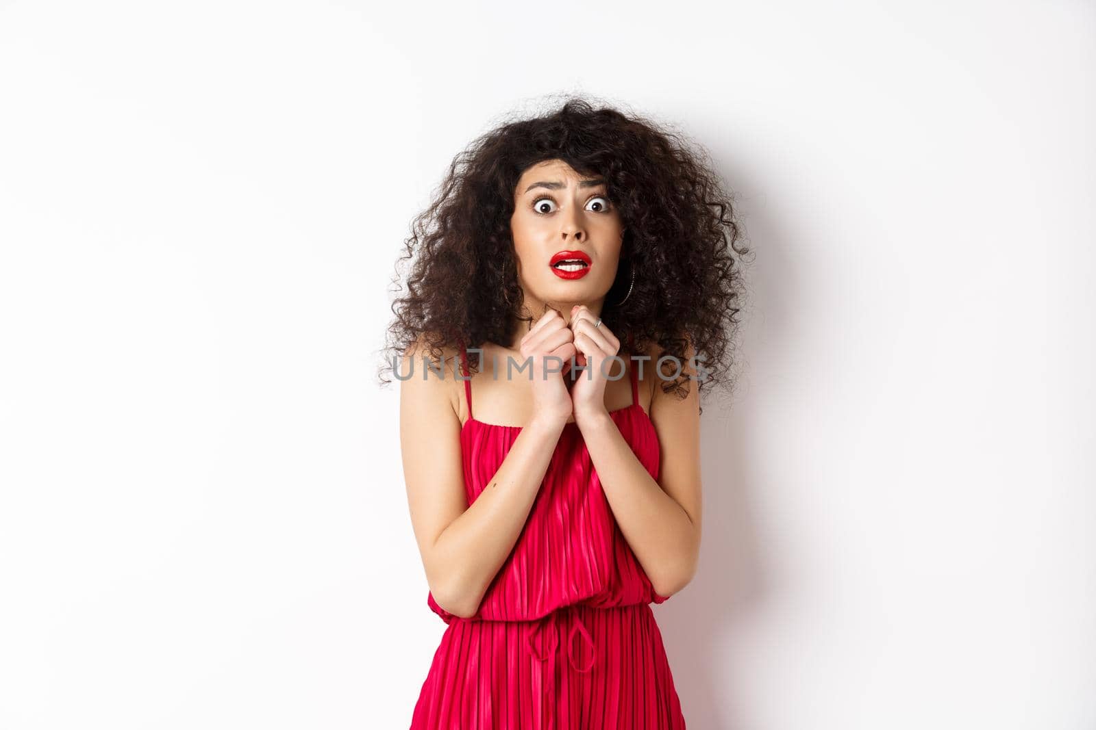 Scared caucasian woman trembling from fear, wearing red dress and staring anxious at camera, standing over white background.