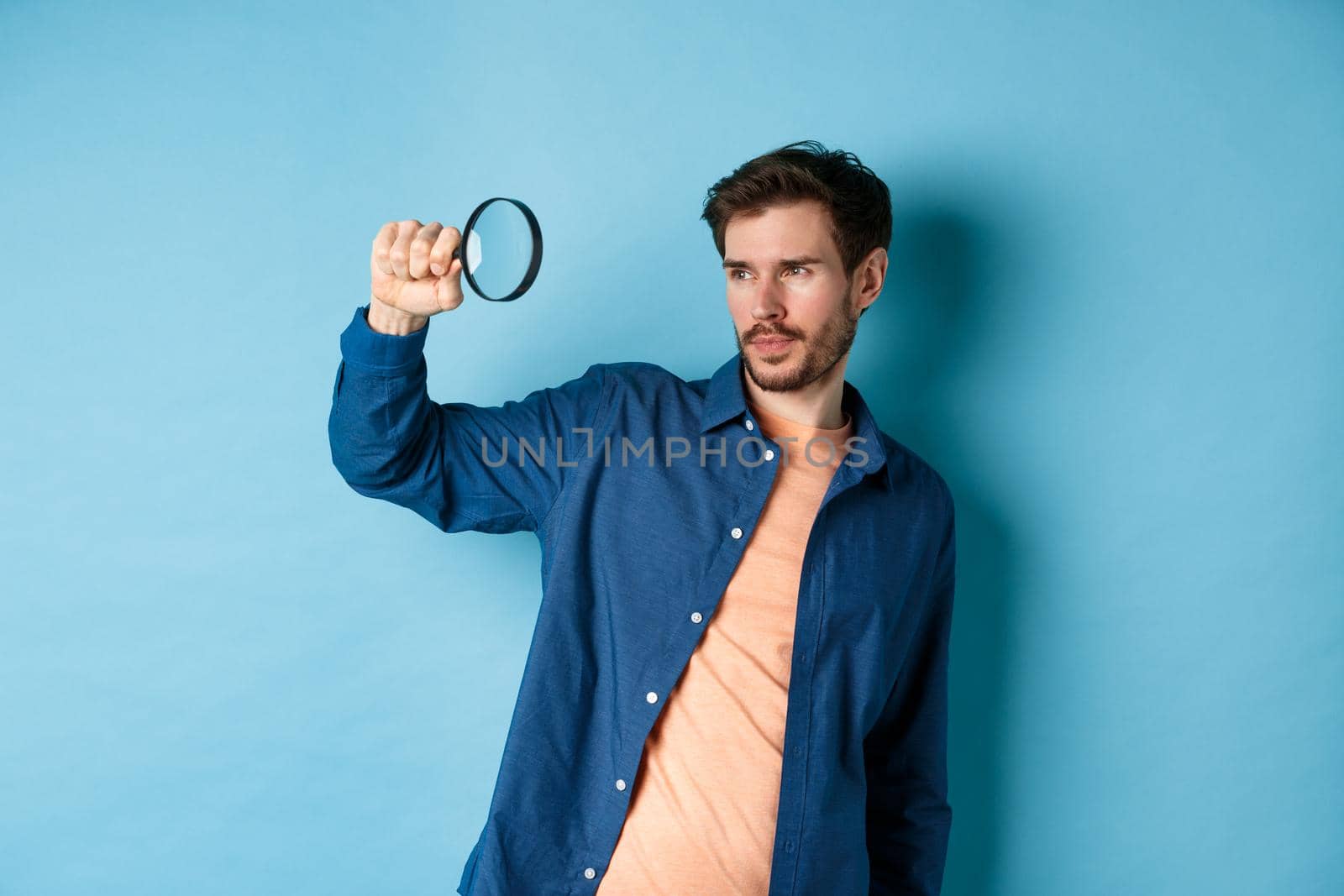 Image of young man searching for something, looking at empty space with magnifying glass, standing on blue background by Benzoix