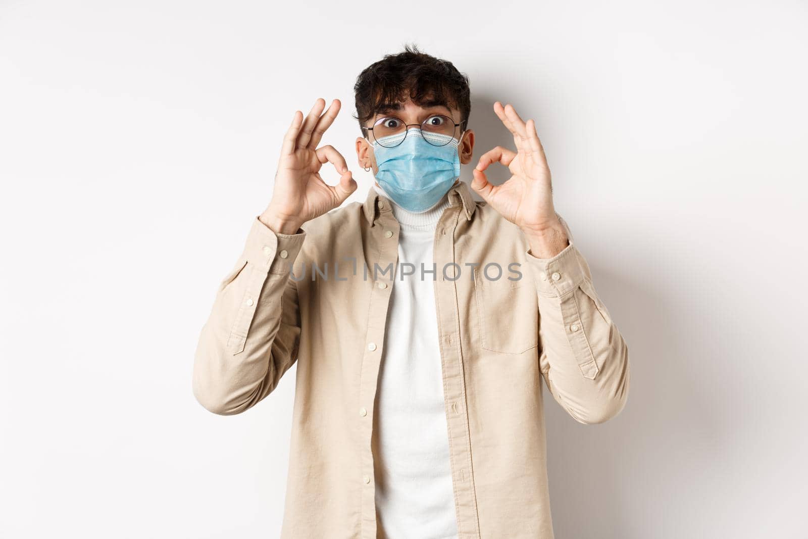 Covid-19, health and real people concept. Excited and impressed guy in sterile face mask showing OK signs in approval, praise cool thing, standing amused on white background.