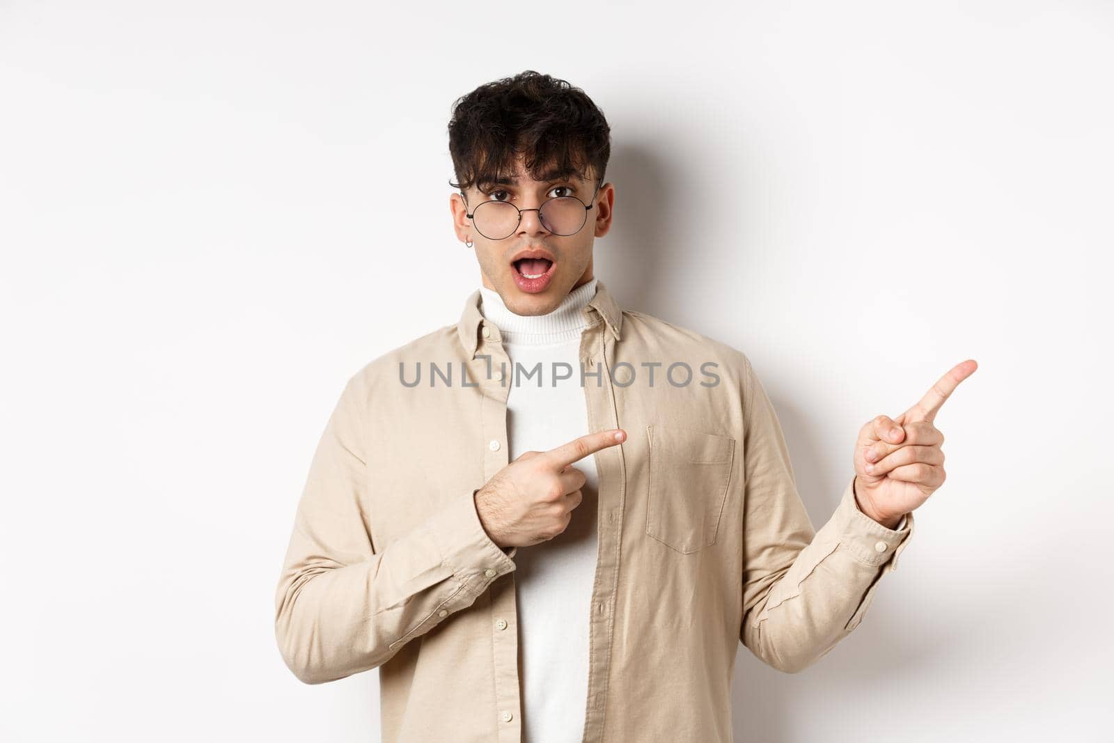 Shocked young man in glasses pointing fingers right at empty space, asking about advertisement, standing on white background.