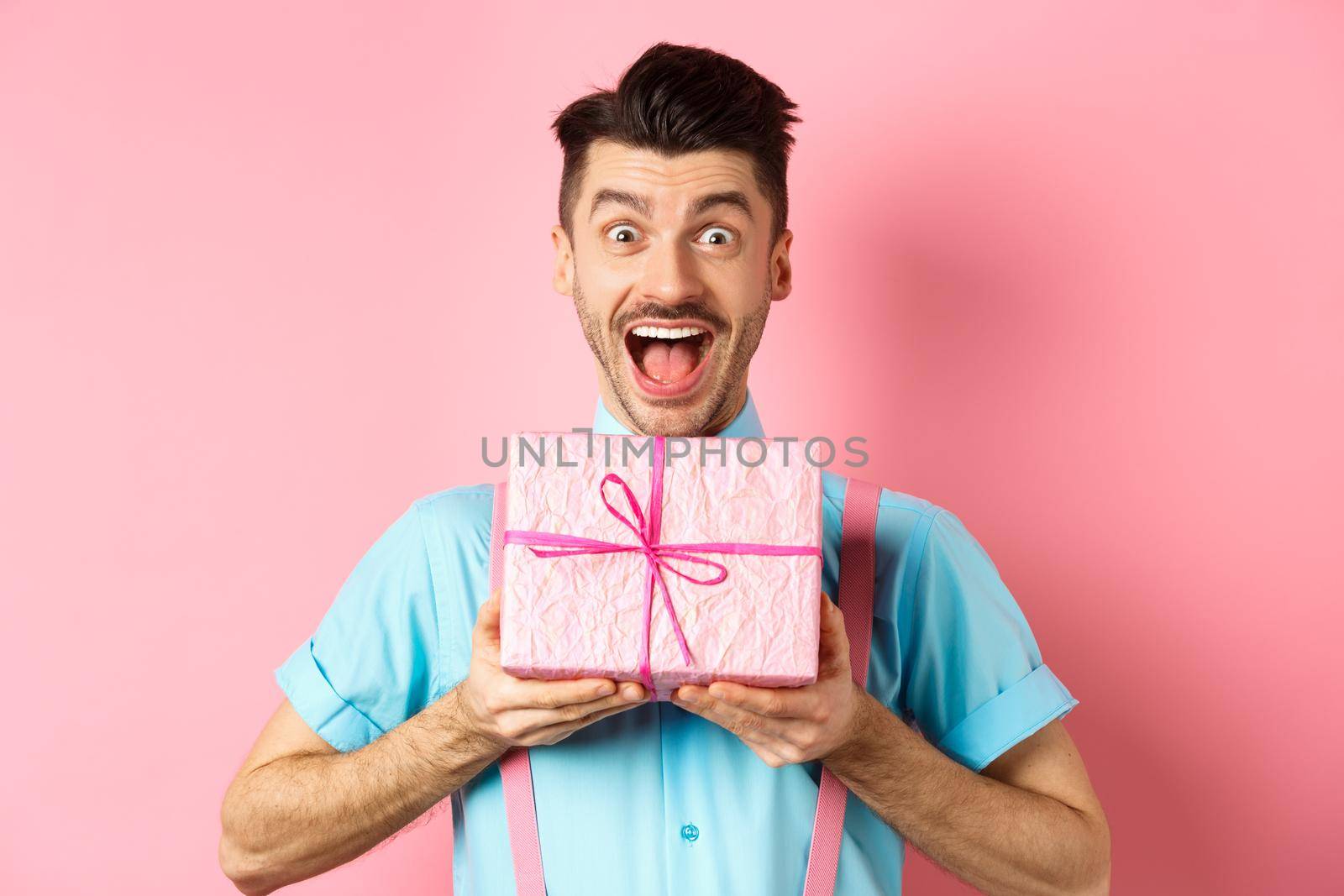Holidays and celebration concept. Excited and surprised guy celebrating birthday, receiving gift and cheering, smiling happy at camera, standing over pink background.